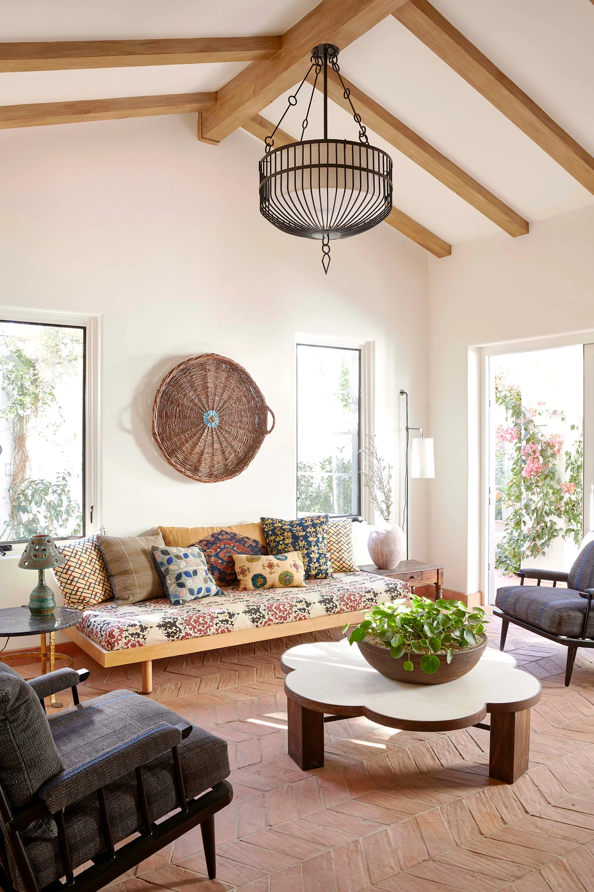 A sunlit living room features terracotta herringbone tile floors, a scalloped wood coffee table, and exposed ceiling beams with a daybed sofa.