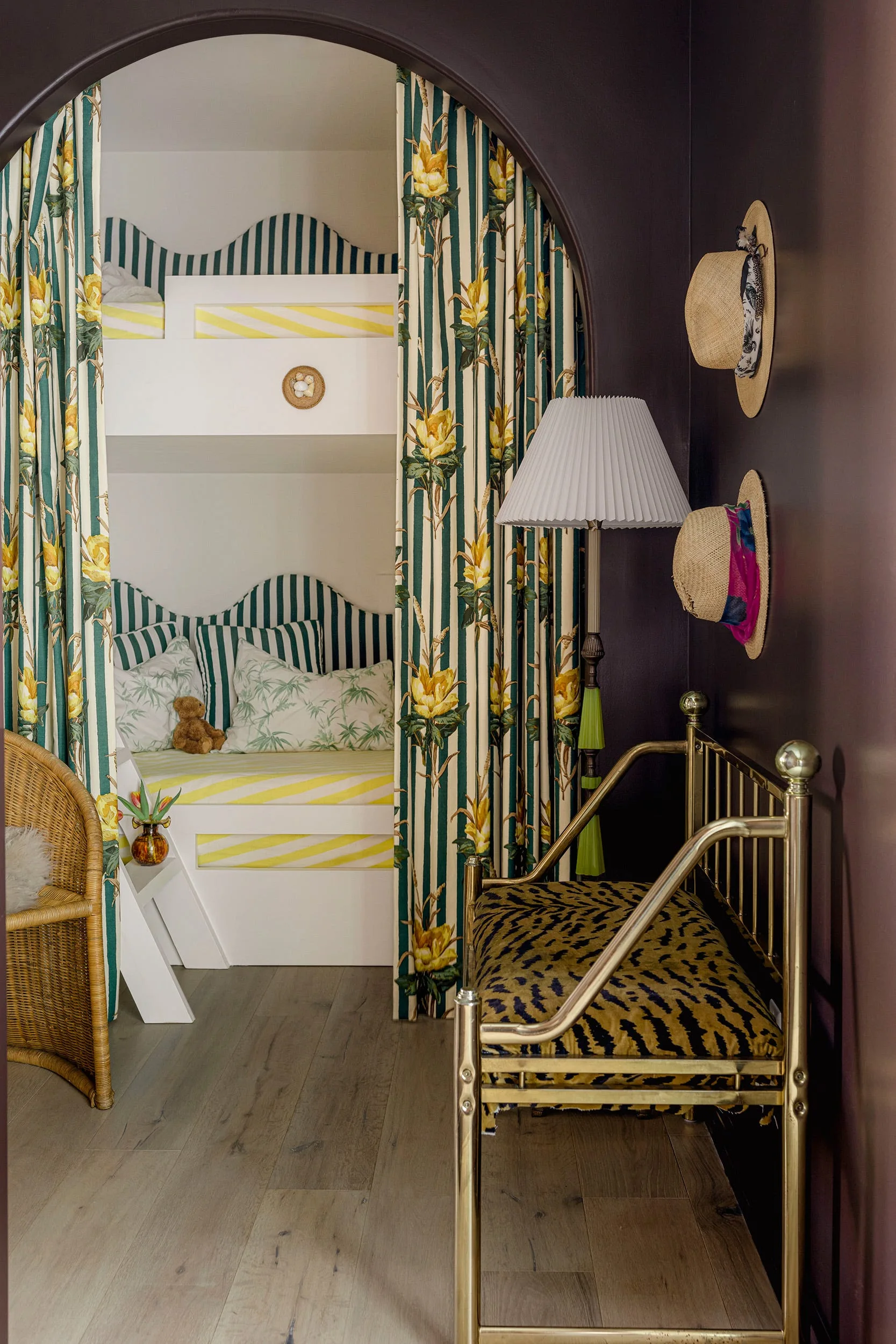 A dark purple archway frames a children's bedroom nook with a white bunk bed, floral curtains, and a patterned brass bench in the foreground.