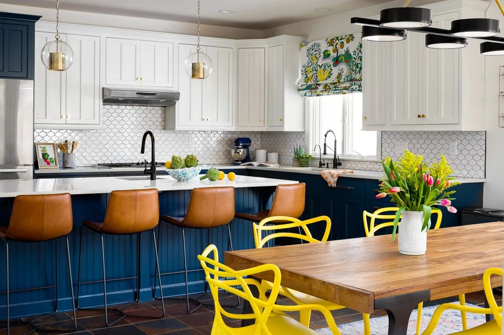 A transitional kitchen features a navy blue island with tan leather barstools, white upper cabinetry, and a wooden dining table with bright yellow chairs.