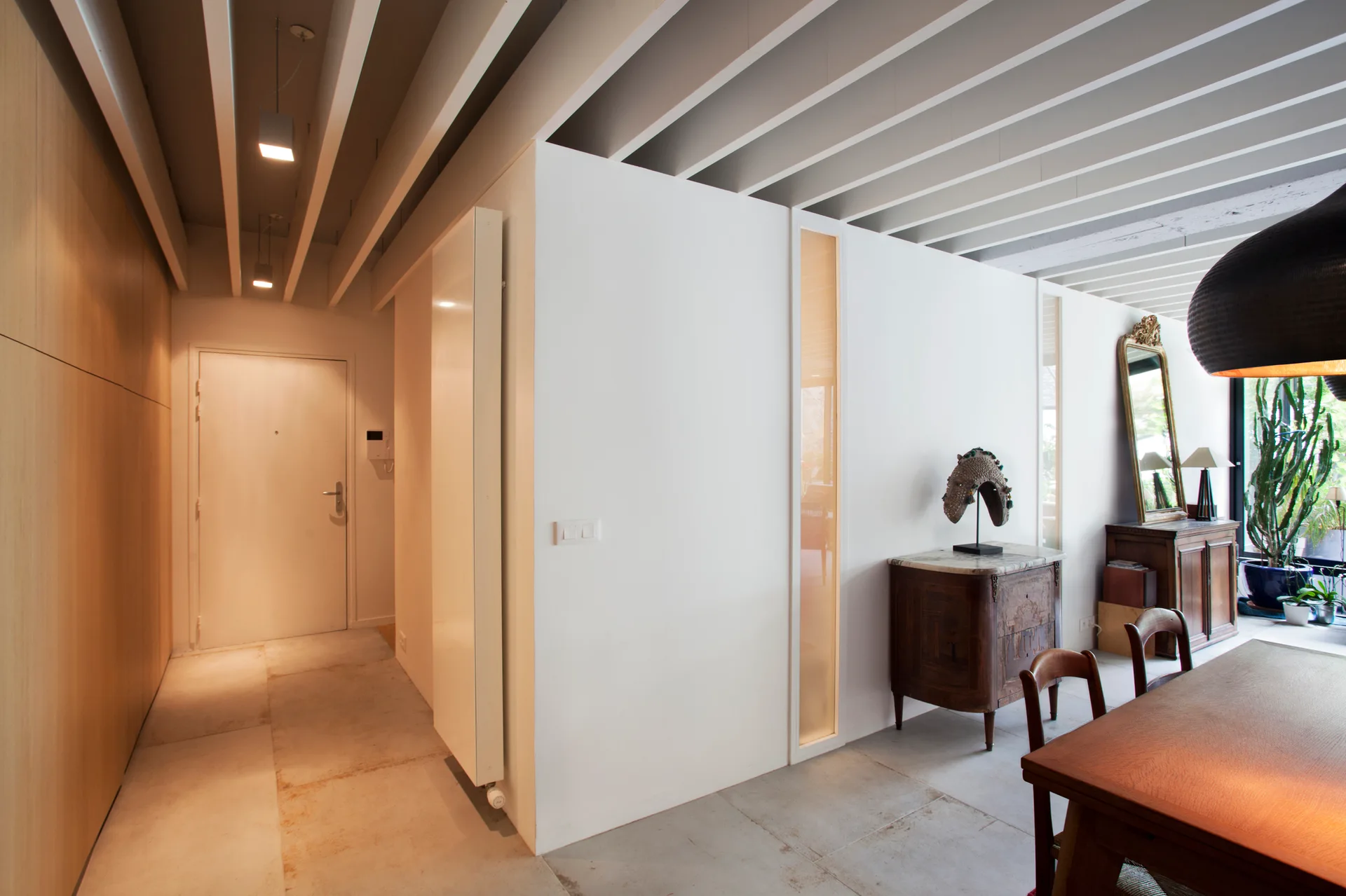 A modern dining room and entry hallway featuring a white slatted ceiling, a wooden dining table, a white partition wall, and an antique sideboard.