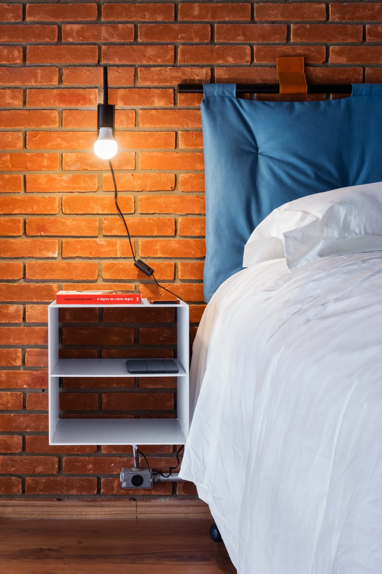 A blue cushioned headboard hangs against an exposed brick wall next to a floating white open-shelf nightstand and a glowing black wall lamp.