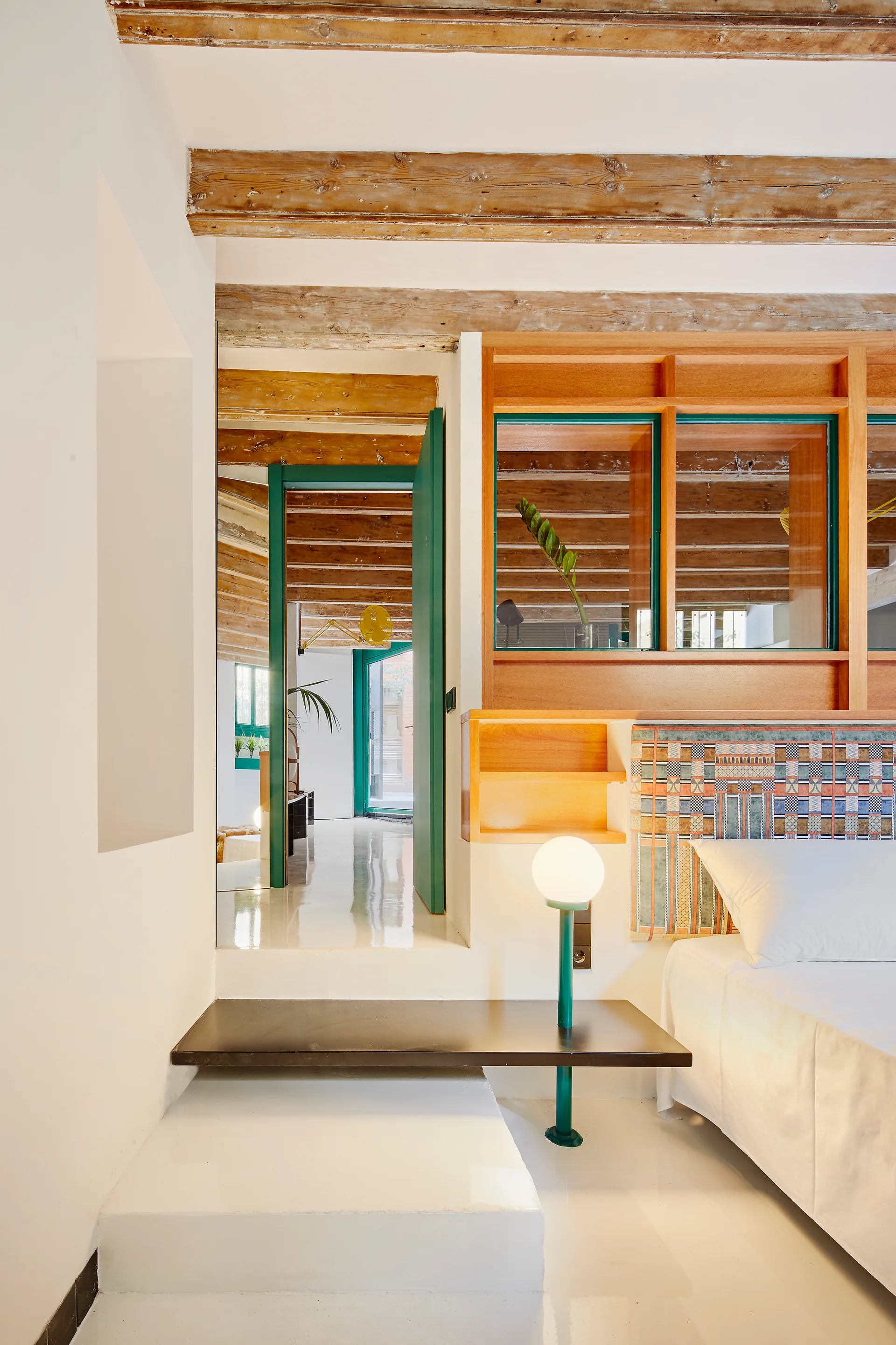 A modern bedroom features a bed with a patterned headboard, exposed wood ceiling beams, a green globe lamp, and a green-framed doorway leading up a step.