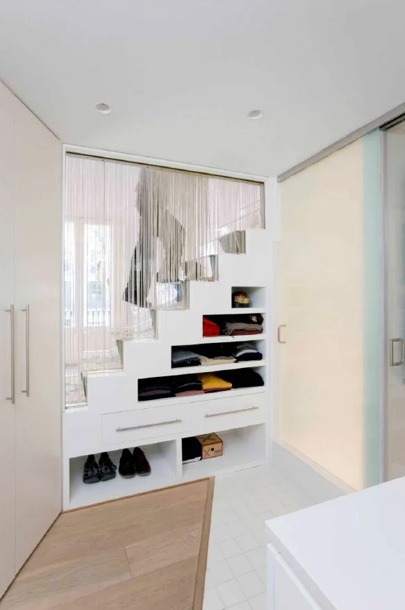 A modern white staircase features built-in drawers, open clothing cubbies, and shoe shelves underneath, positioned near a frosted glass sliding door.