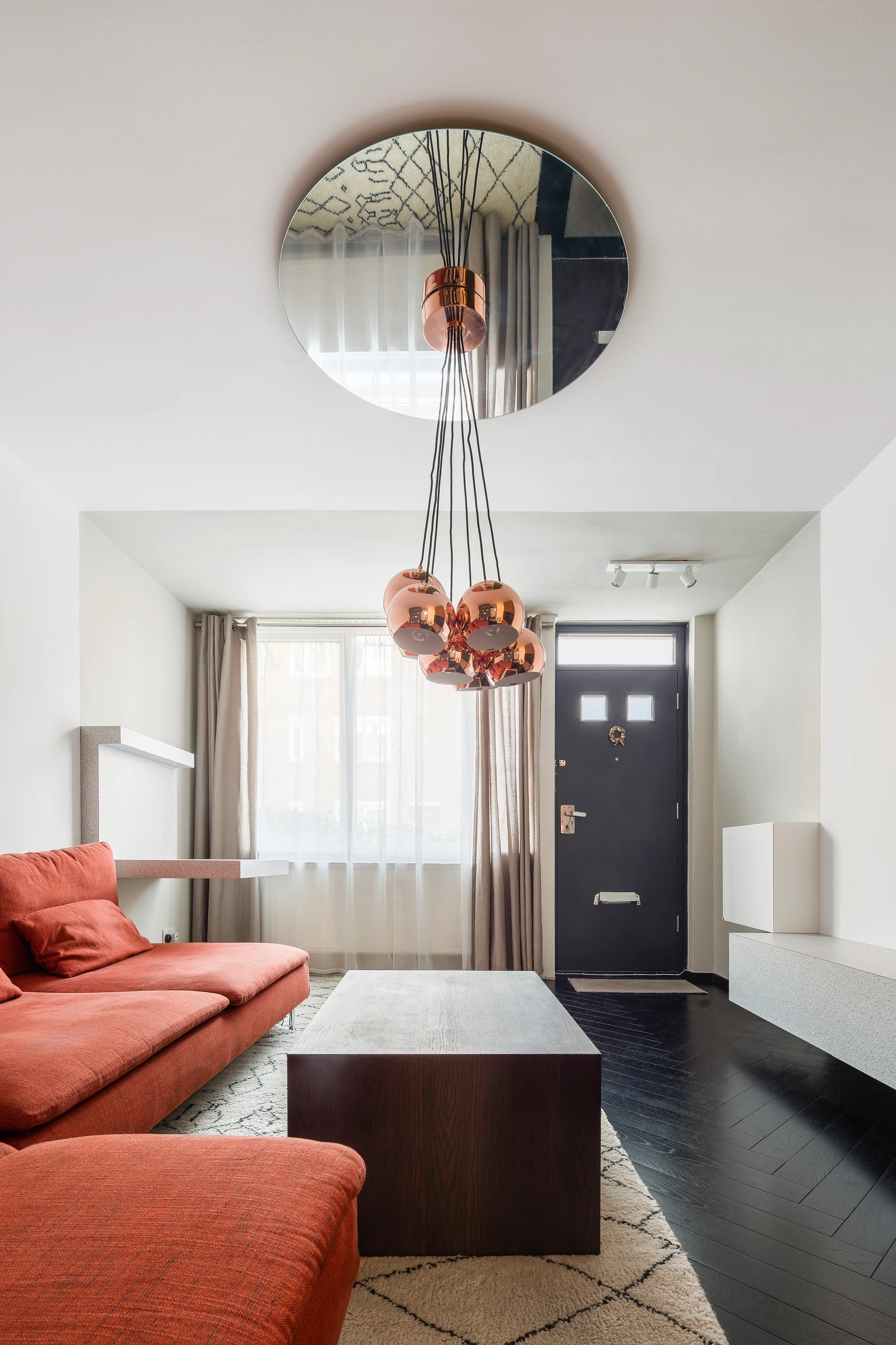 A living room features a red sectional sofa, dark wood coffee table, and a circular ceiling mirror reflecting copper globe pendant lights.