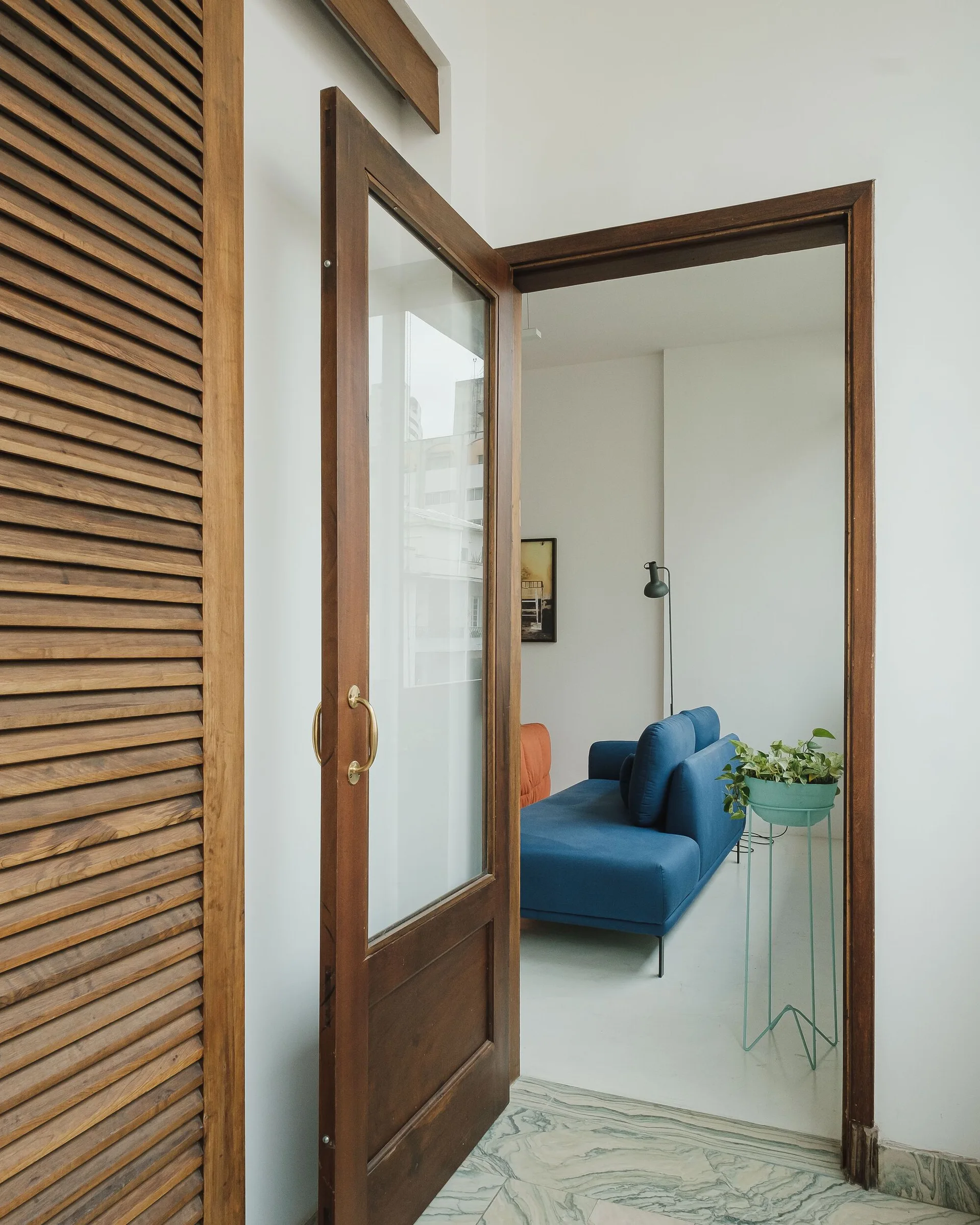 A view through an open dark wood door with glass panes reveals a bright living room featuring a blue sofa, white walls, and a teal plant stand.