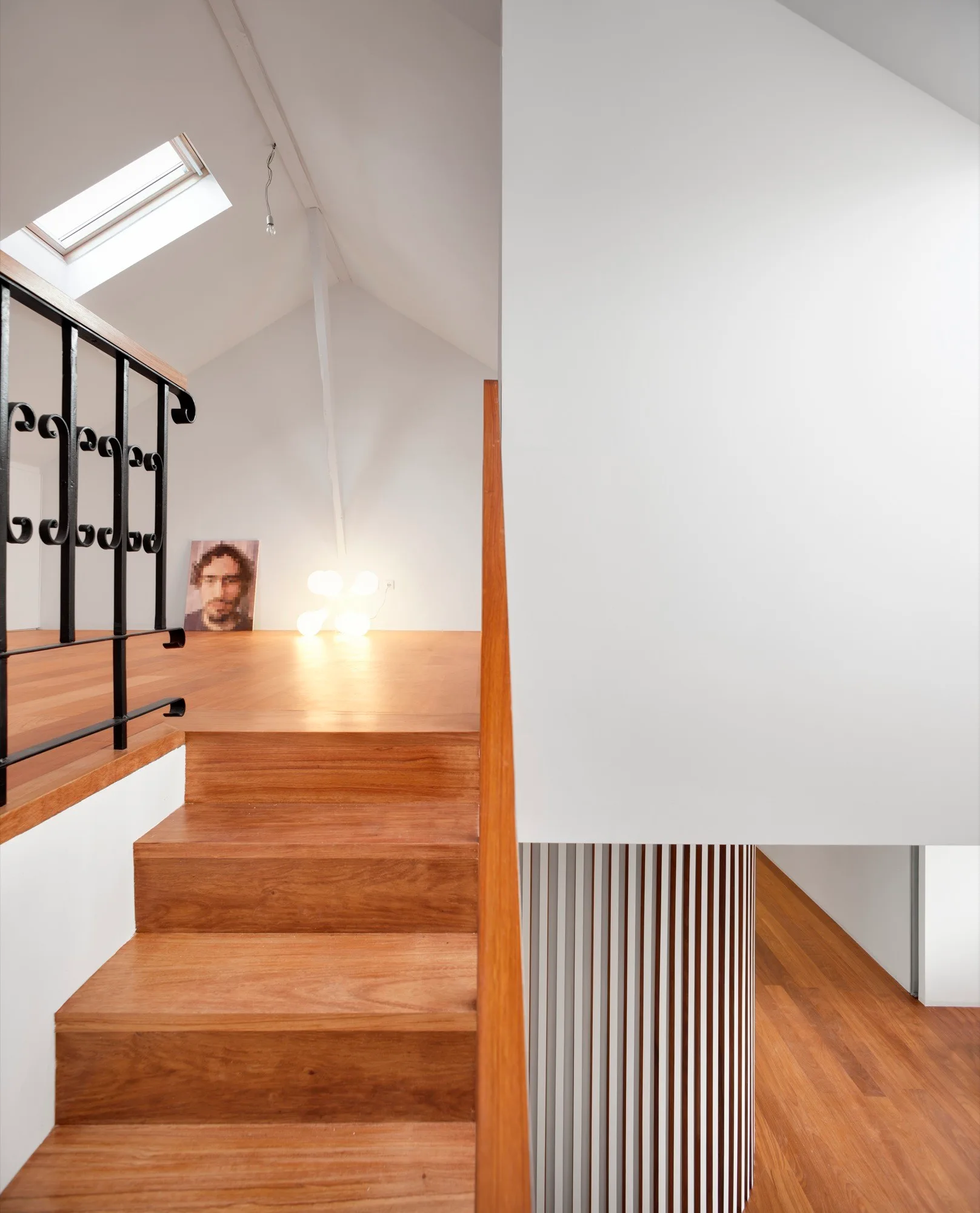 A wooden staircase leads to a minimalist loft with a sloped ceiling, skylight, black metal railing, and white walls.