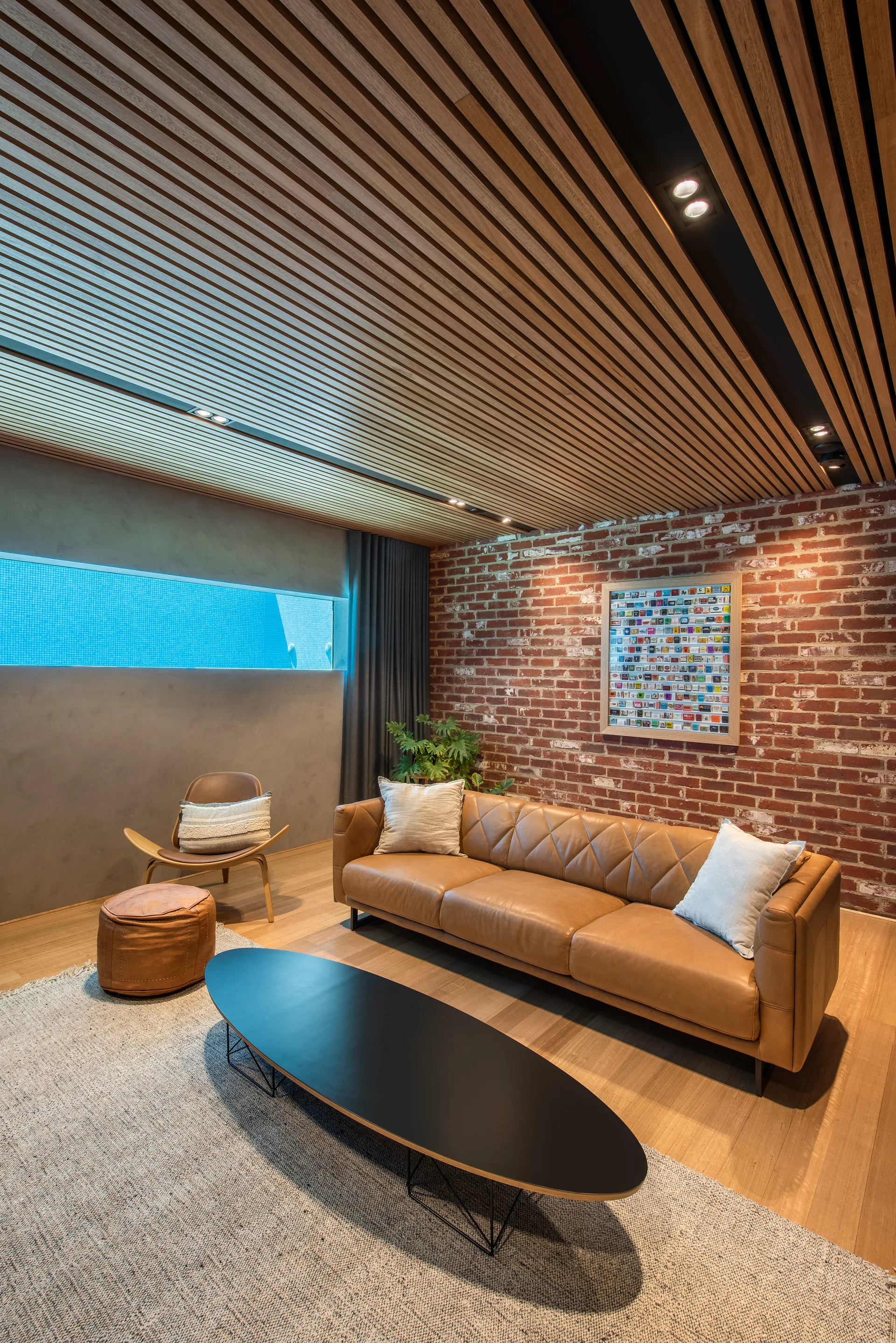A modern living room featuring a tan leather sofa, a black oval coffee table, a brick accent wall, and a linear slatted wood ceiling.