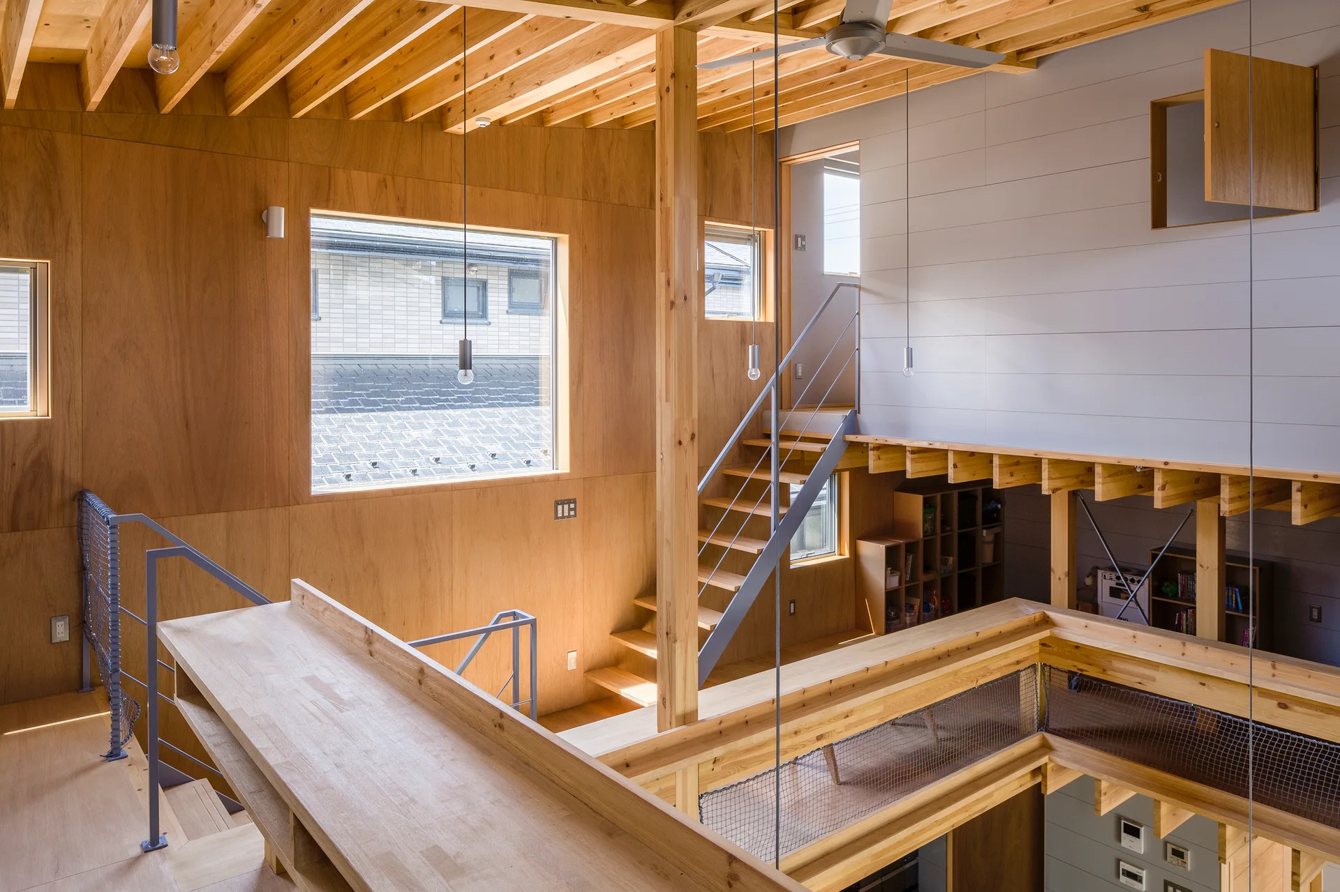 A modern multi-level residential interior featuring warm plywood walls, a central wooden staircase with metal railings, and exposed timber ceiling beams.