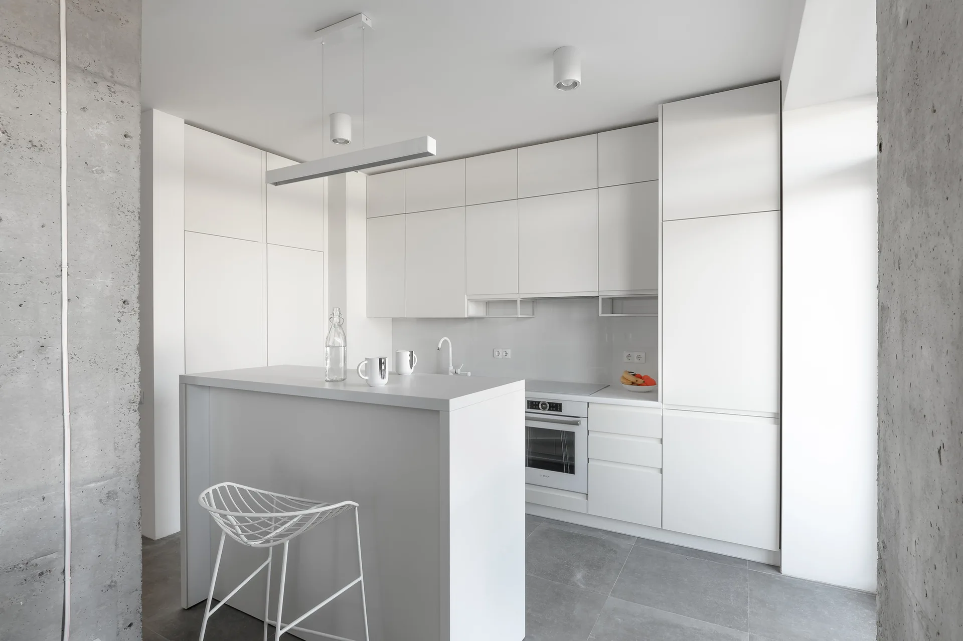 A minimalist white kitchen featuring flat-panel handleless cabinets, a matching island with a white wire stool, and an exposed concrete pillar.
