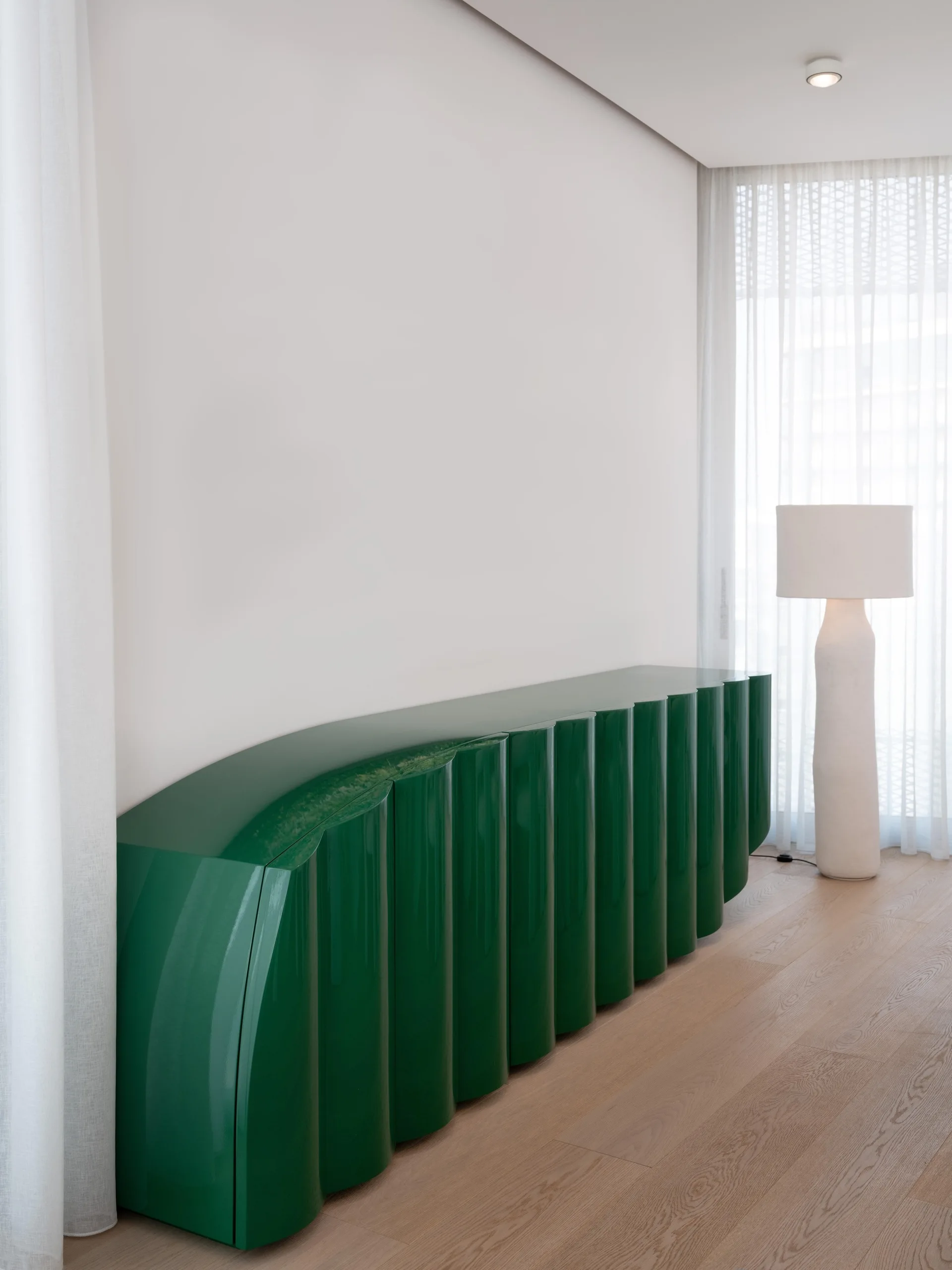 A glossy green fluted credenza stands against a white wall next to a tall white floor lamp and sheer curtains.