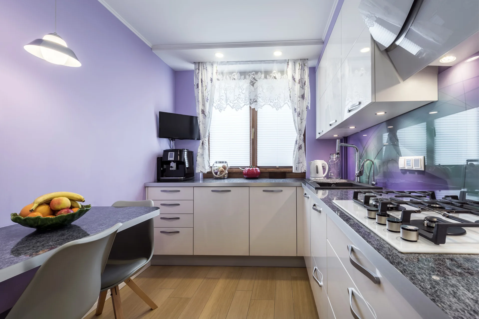 A compact kitchen featuring lavender walls, glossy white cabinets, a dark speckled stone countertop, and a small dining table with grey chairs.