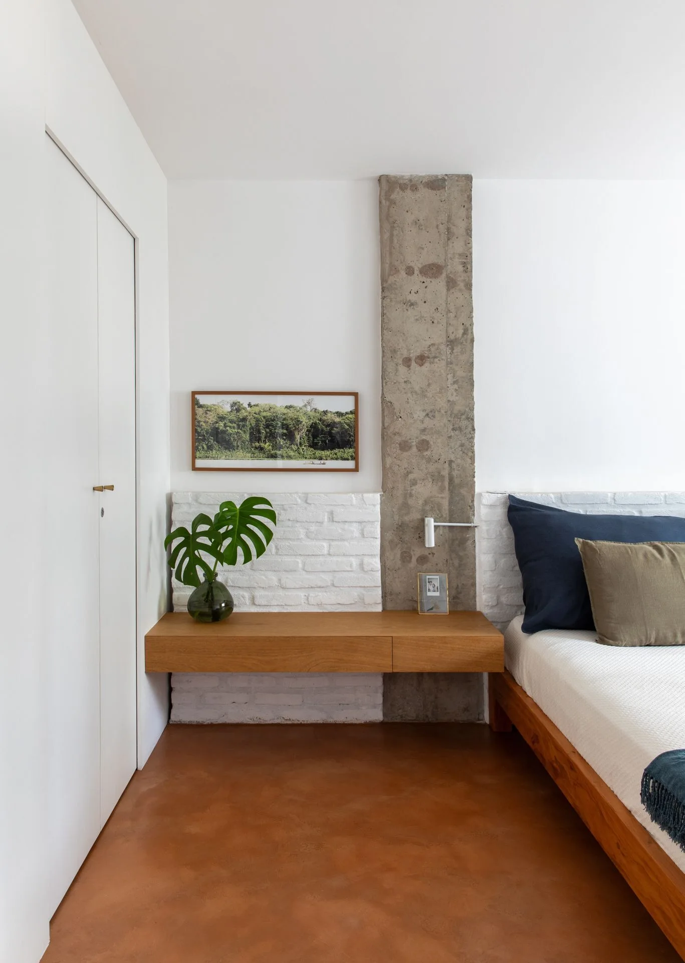 A minimalist bedroom featuring a floating wood bedside shelf, an exposed concrete column, white painted brick, and matte terracotta floors.