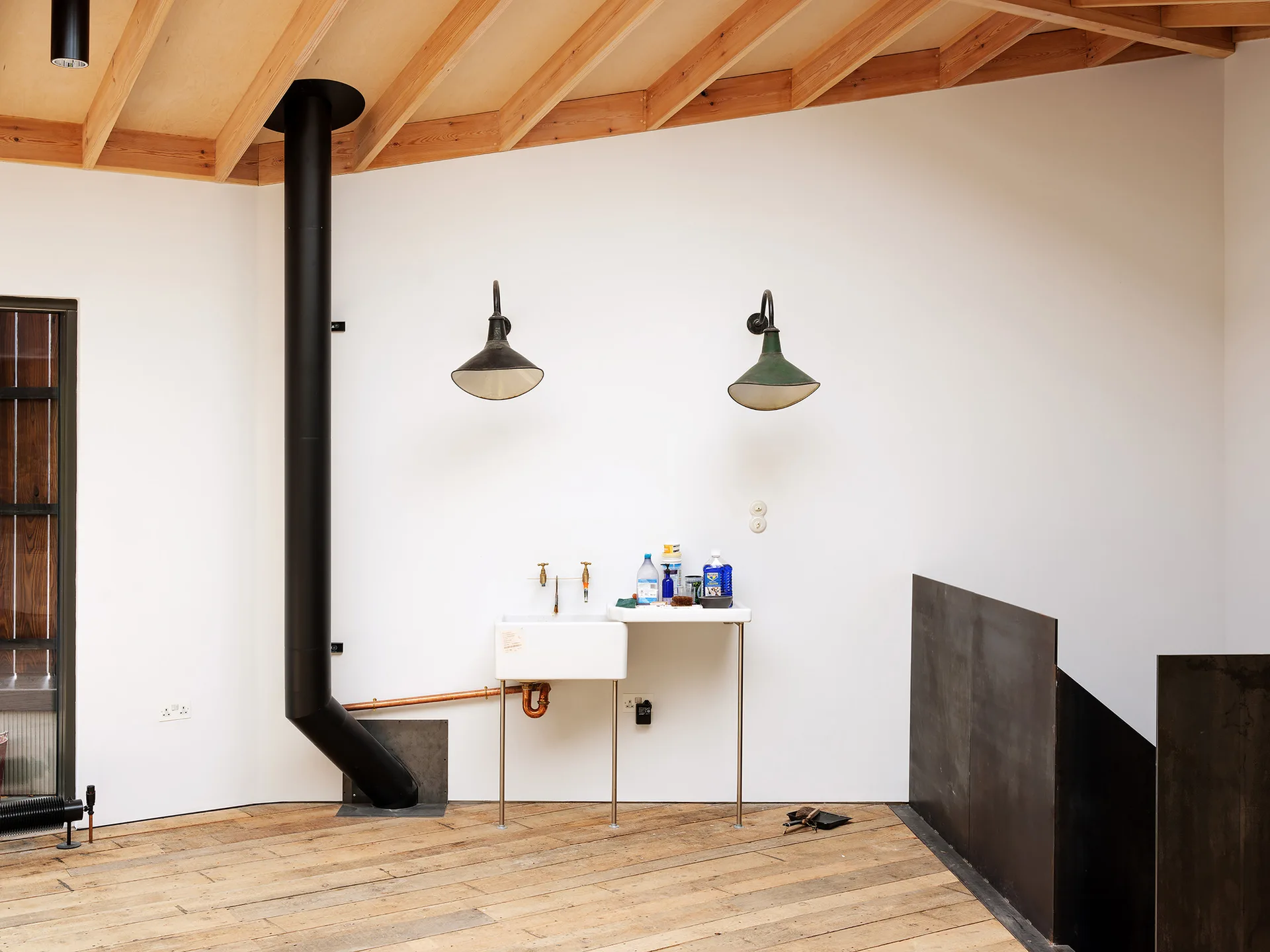 A white utility sink with brass faucets stands beneath two dark sconces in a room with a black metal flue and an exposed wood ceiling.