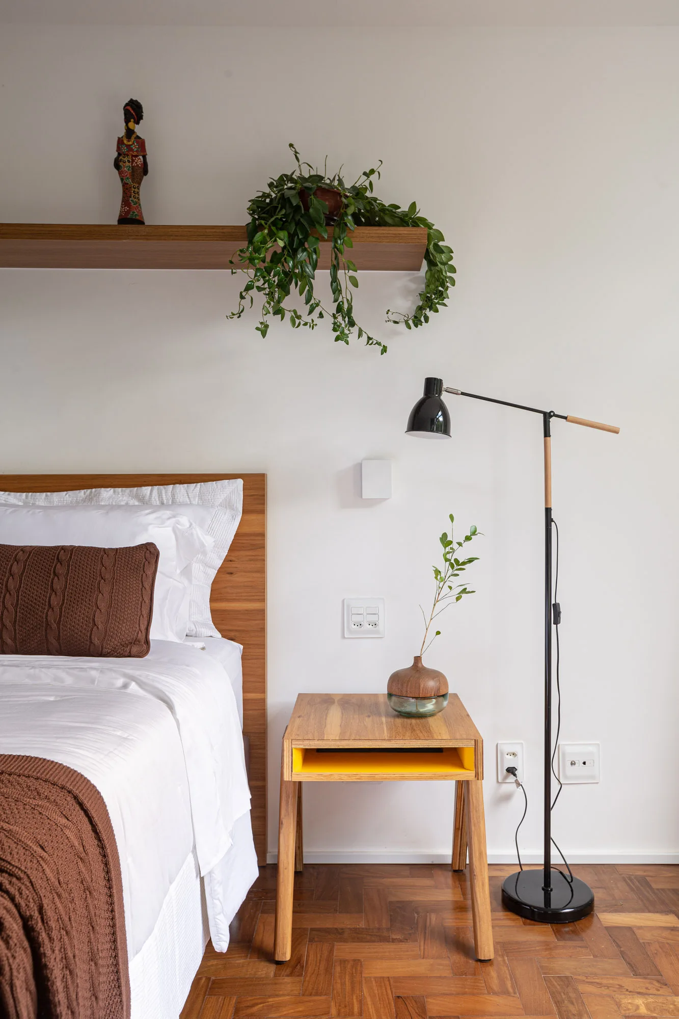 A minimalist bedroom features a wooden bed with white linens, a wooden nightstand with a yellow compartment, a black floor lamp, and a floating shelf.