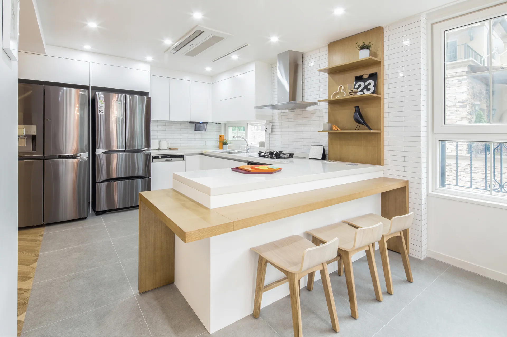 A modern white kitchen features a central island with an integrated wood-wrapped breakfast bar, wooden bar stools, and white subway tile backsplash.