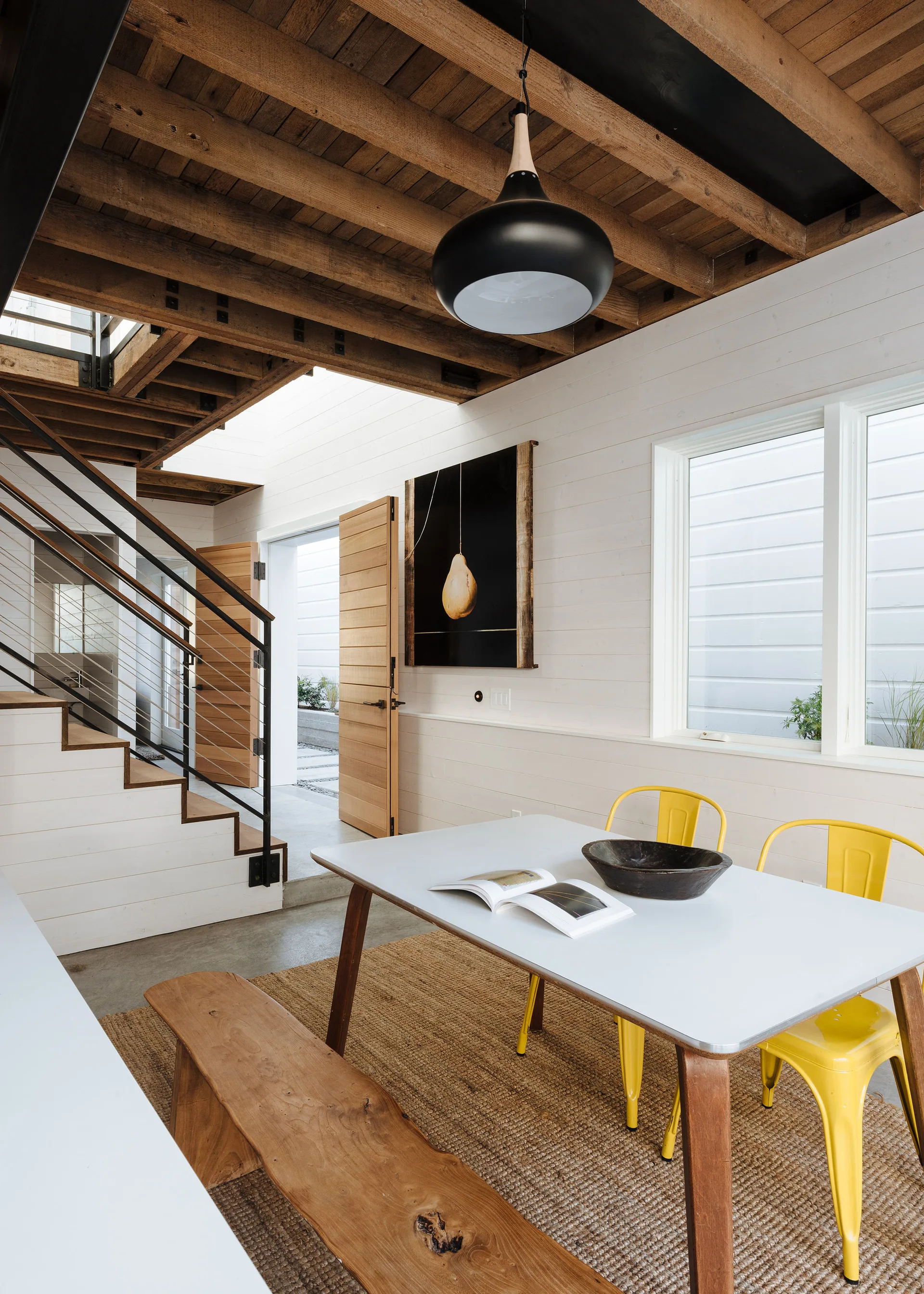 A bright dining space features a white table paired with vibrant yellow chairs, a rustic wooden bench, and exposed wooden ceiling beams.