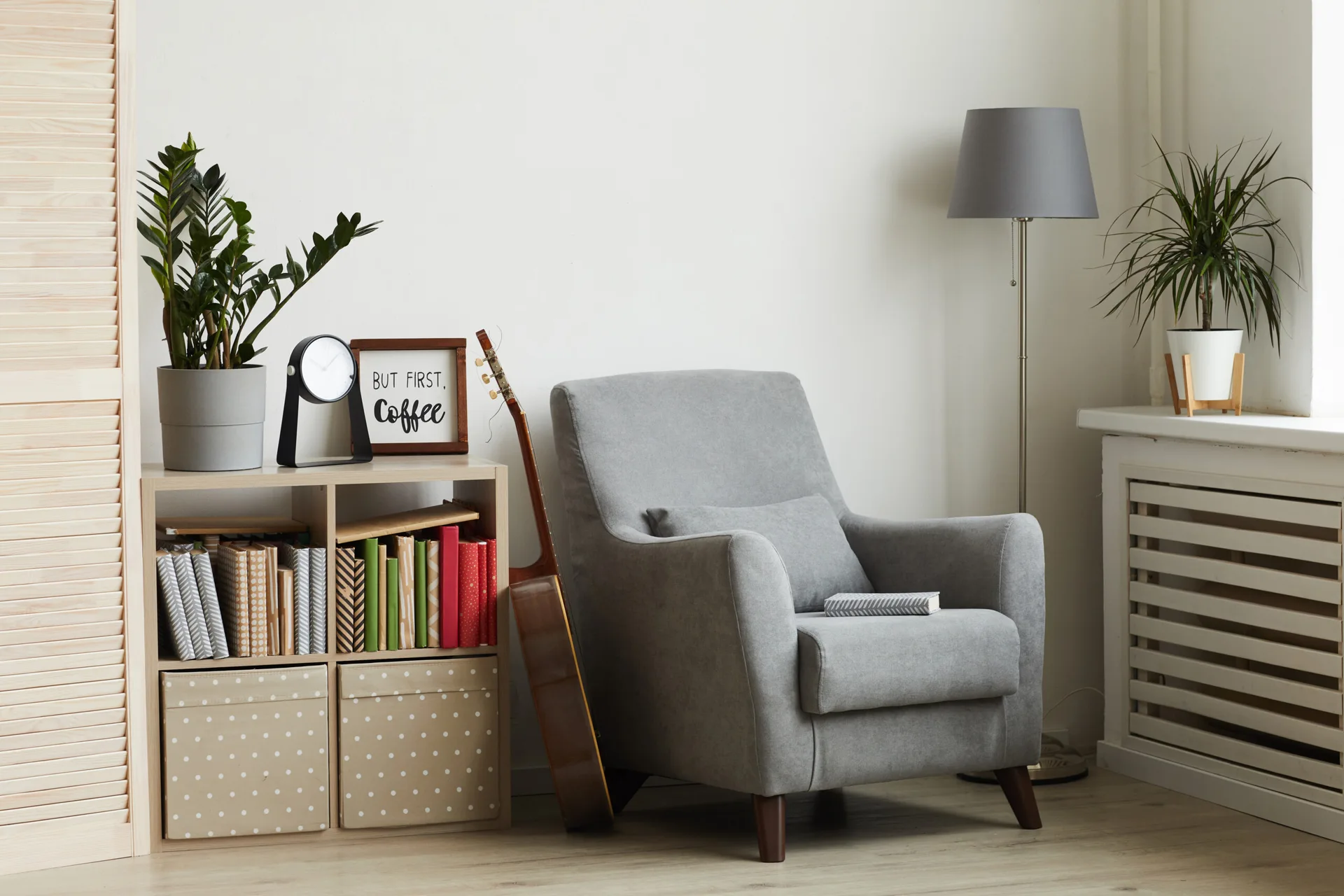 A gray fabric armchair stands in a bright room corner next to a light wood cubby bookshelf holding colorful books and a leaning acoustic guitar.