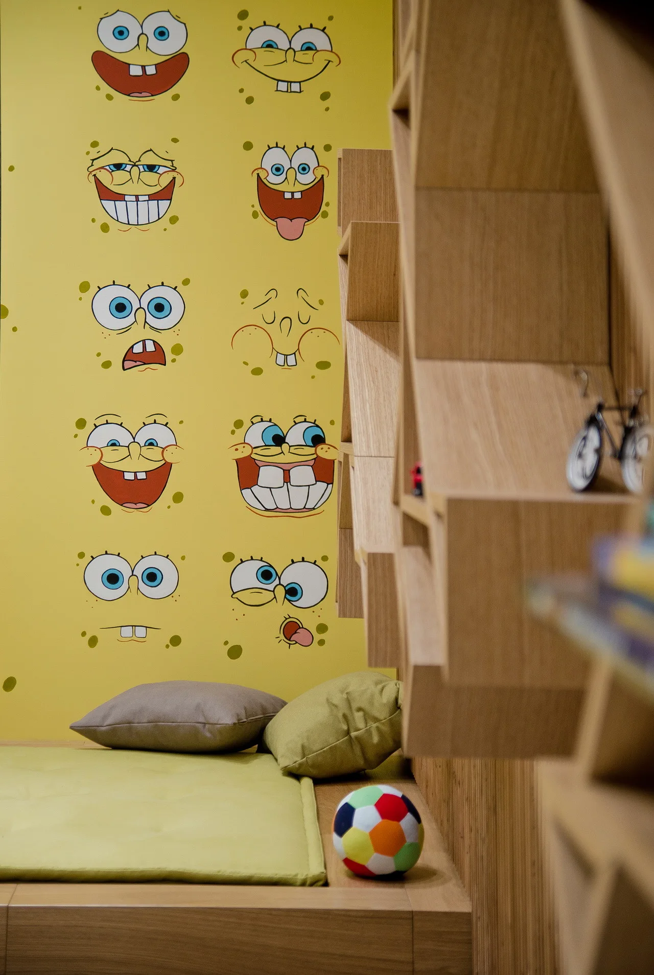 A children's bedroom features bright yellow character wallpaper behind a platform daybed and a custom geometric wooden shelving unit.
