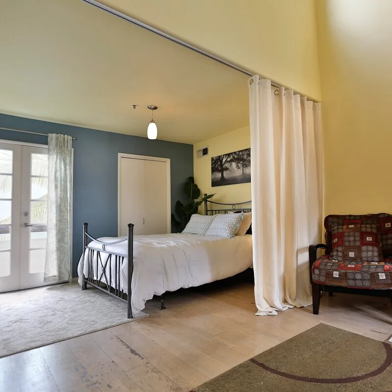 A bedroom with yellow walls, a blue-grey accent wall, a black metal bed, and a long cream-colored curtain divider on a ceiling track.