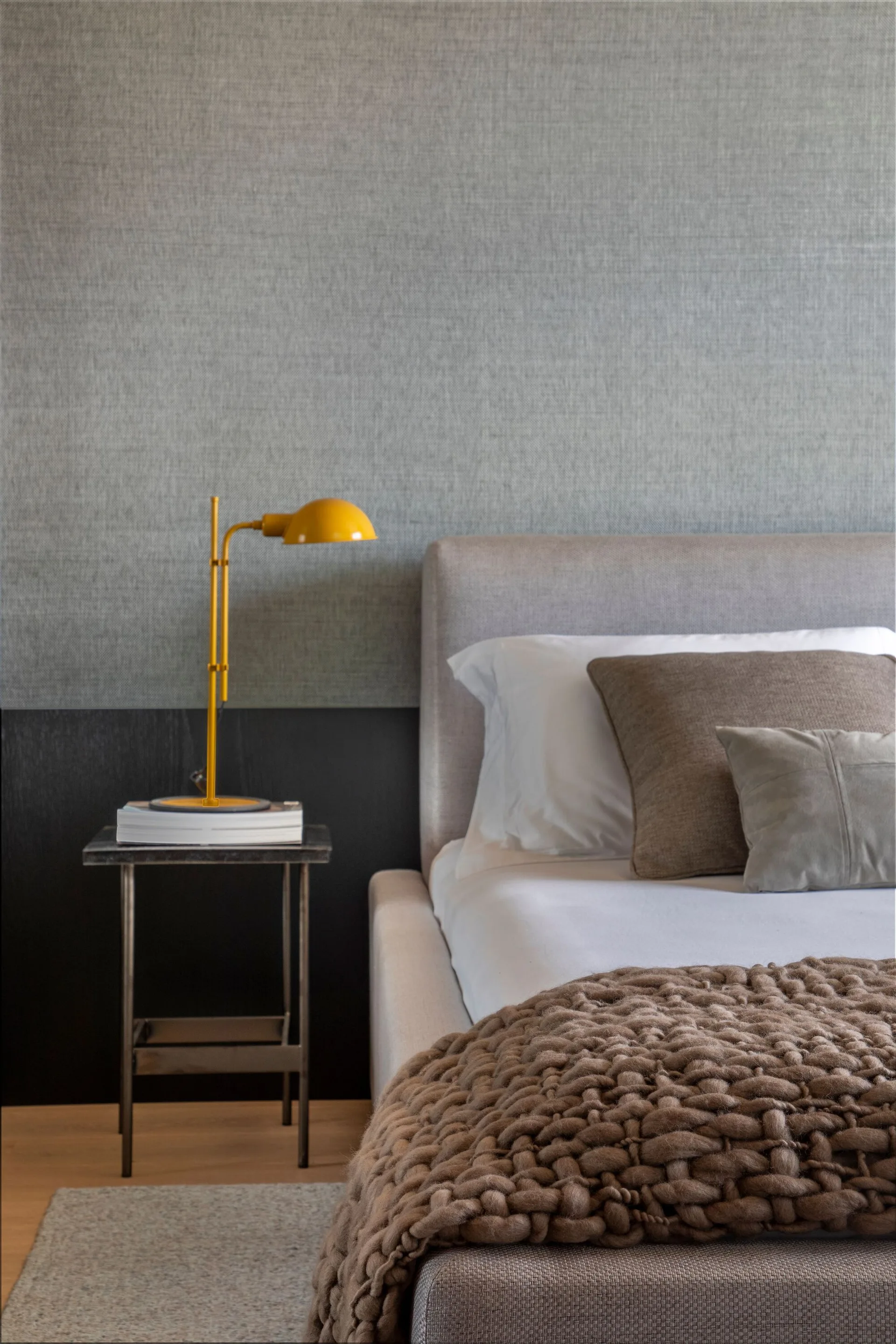 A modern bedroom with a two-tone gray wall, a beige upholstered bed, and a bright yellow task lamp on a metal nightstand.
