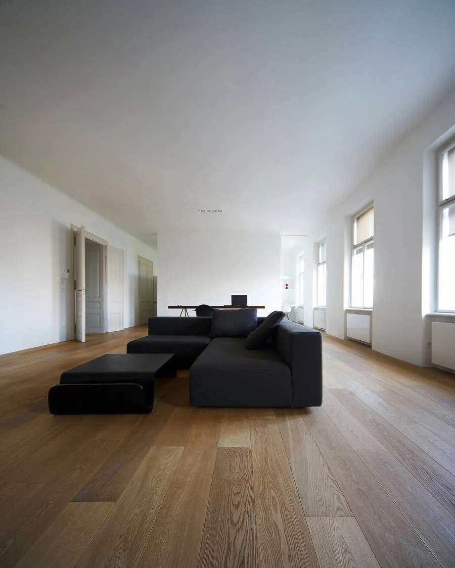 A dark gray sectional sofa and low black coffee table sit on light wood flooring in a minimalist living room with white walls and tall windows.