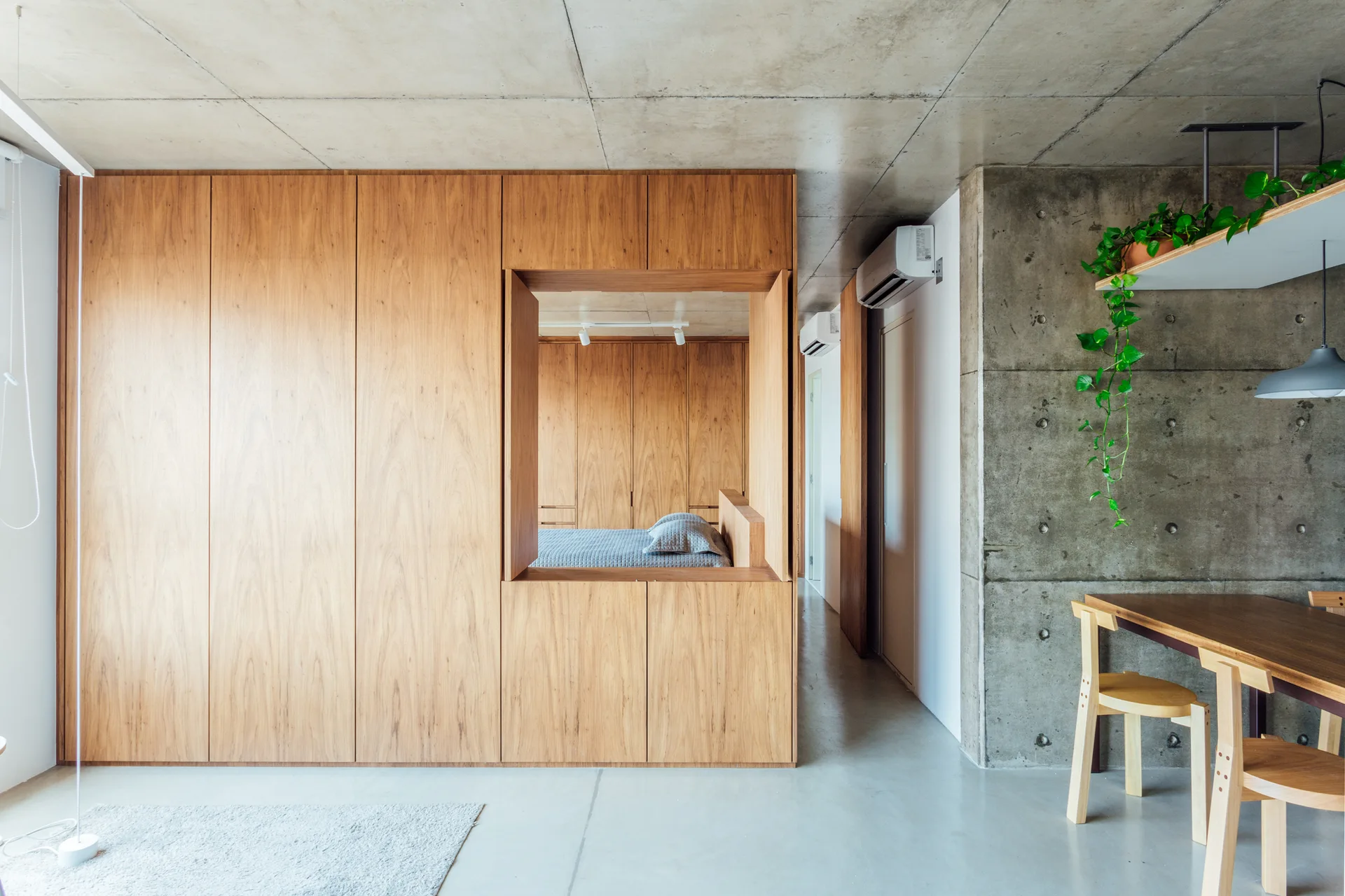 A large wooden storage wall with a central window cutout looking into a bedroom, set against raw concrete walls and flooring.
