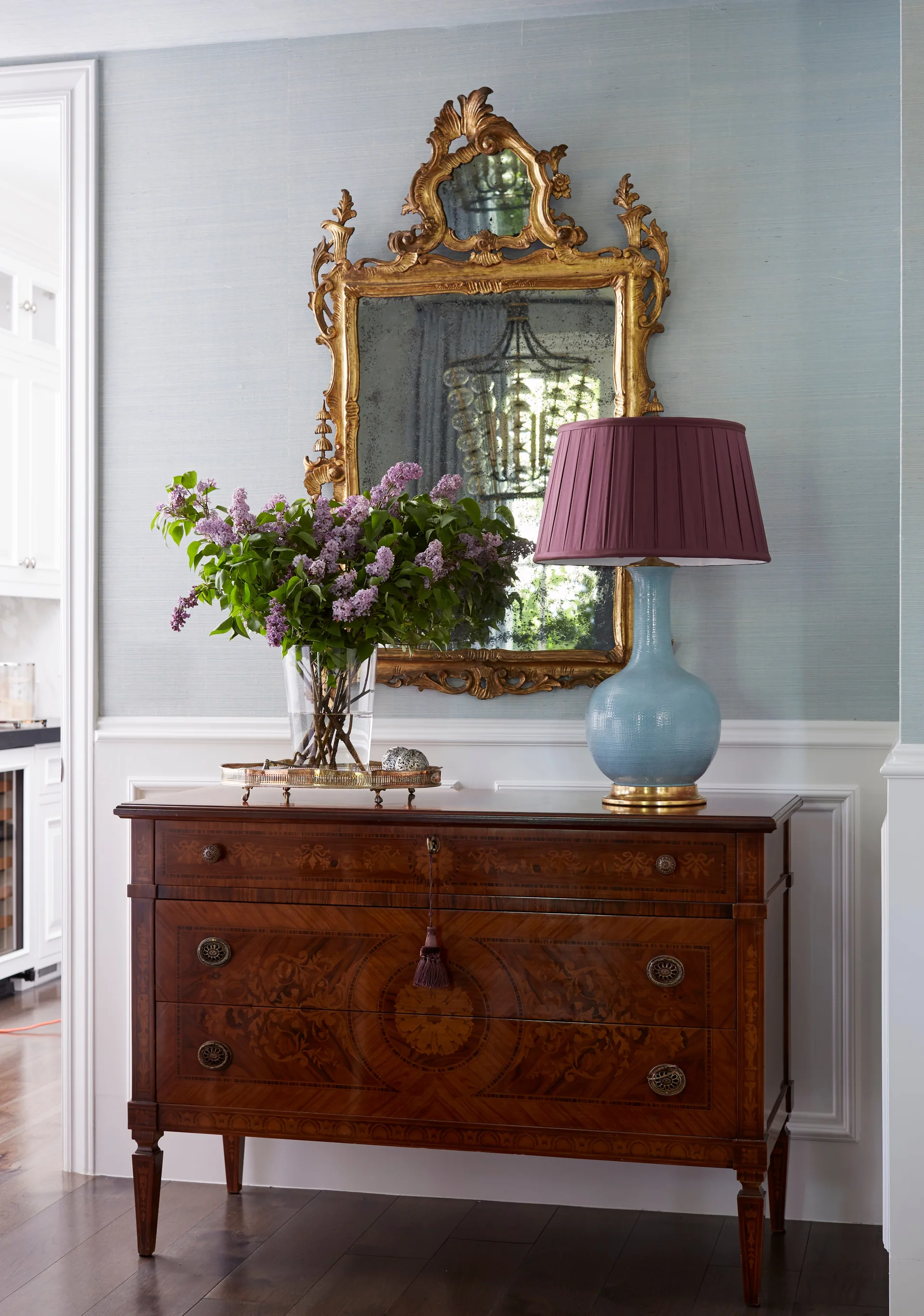 An antique inlaid wood chest sits against blue grasscloth wallpaper, topped with an ornate gold mirror, a blue table lamp, and purple flowers.