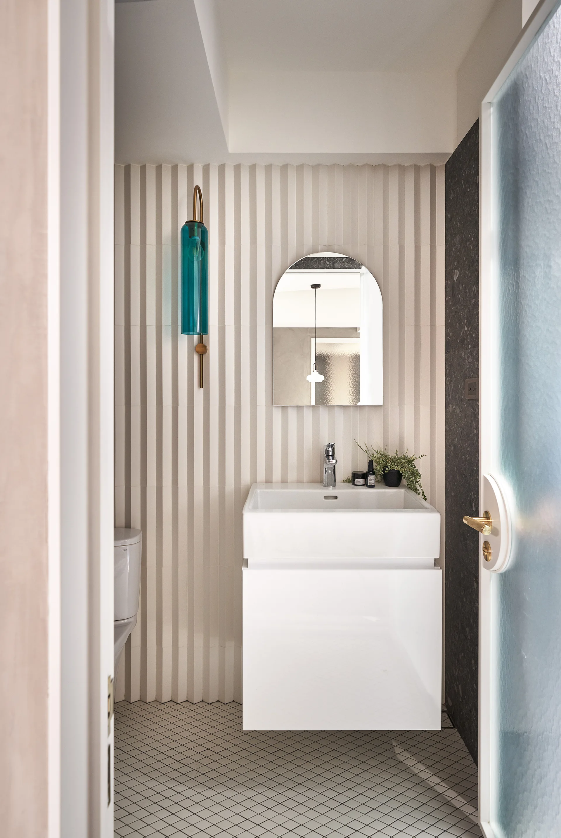 A modern powder room features a white floating vanity under an arched mirror, set against a cream fluted tile wall with a blue glass sconce.