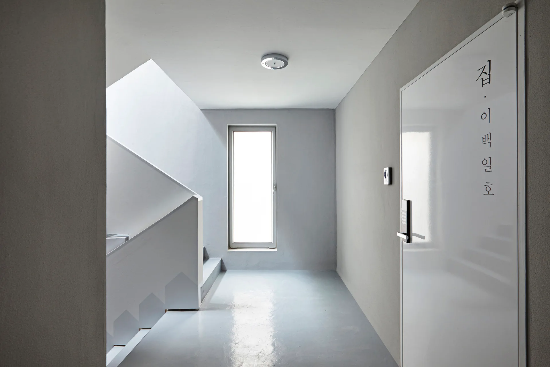 A minimalist grey hallway features a white door on the right, a staircase on the left, and a narrow vertical window reflecting light onto the glossy floor.