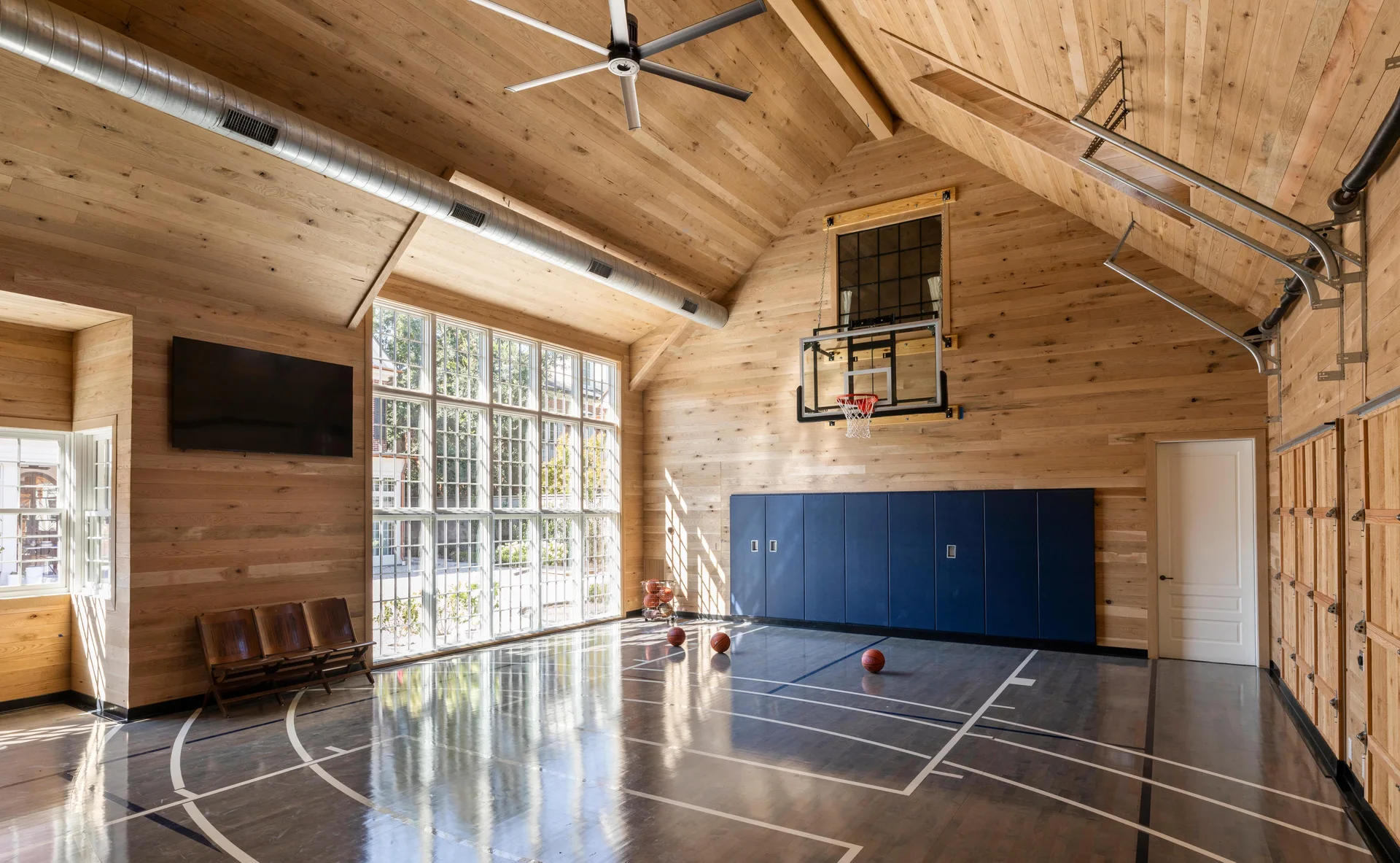 An indoor home basketball court features wood plank walls, a glossy court floor, a basketball hoop, and a large multi-pane window.
