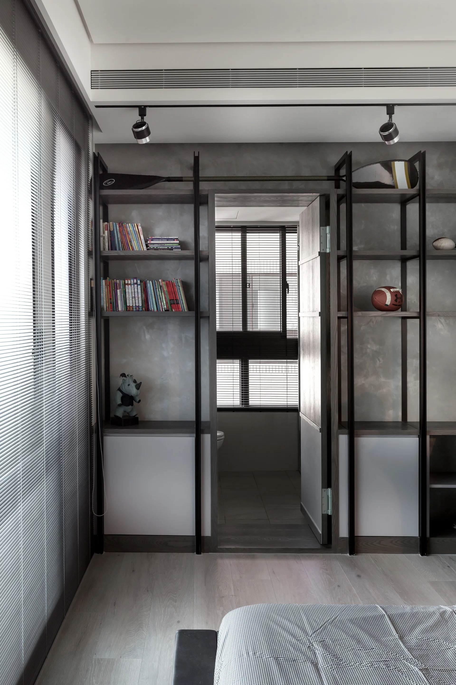 A modern bedroom features symmetrical dark metal shelving built around a doorway, displaying books and sports decor against a textured gray wall.