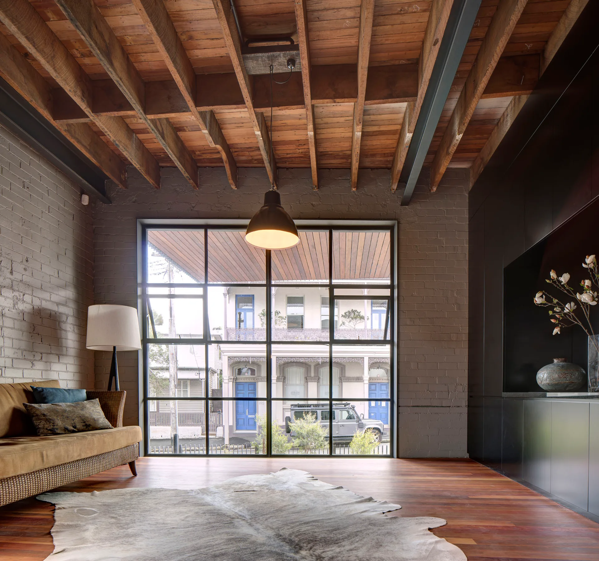 An industrial living room features exposed wooden ceiling joists, a grey painted brick wall, a large grid window, and a brown sofa.