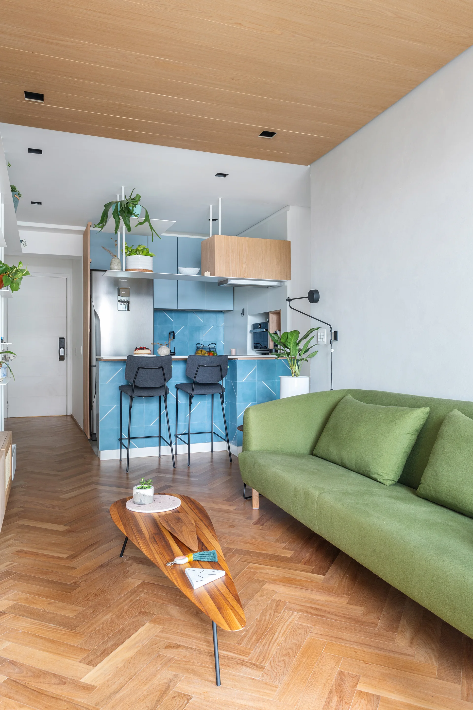 Modern living room featuring a green sofa, teardrop wood coffee table, herringbone flooring, and a kitchen with blue tiled island and cabinets.
