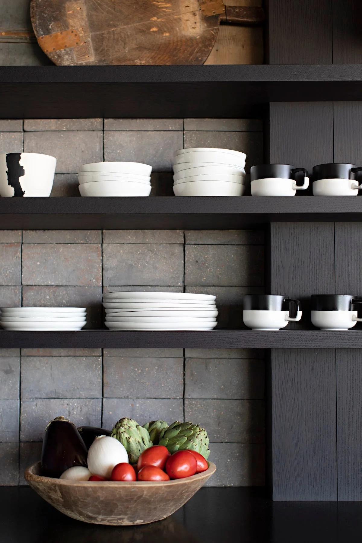 Black wooden shelves display white ceramic plates and two-toned mugs against a grey tiled backsplash with a wooden bowl of fresh vegetables below.