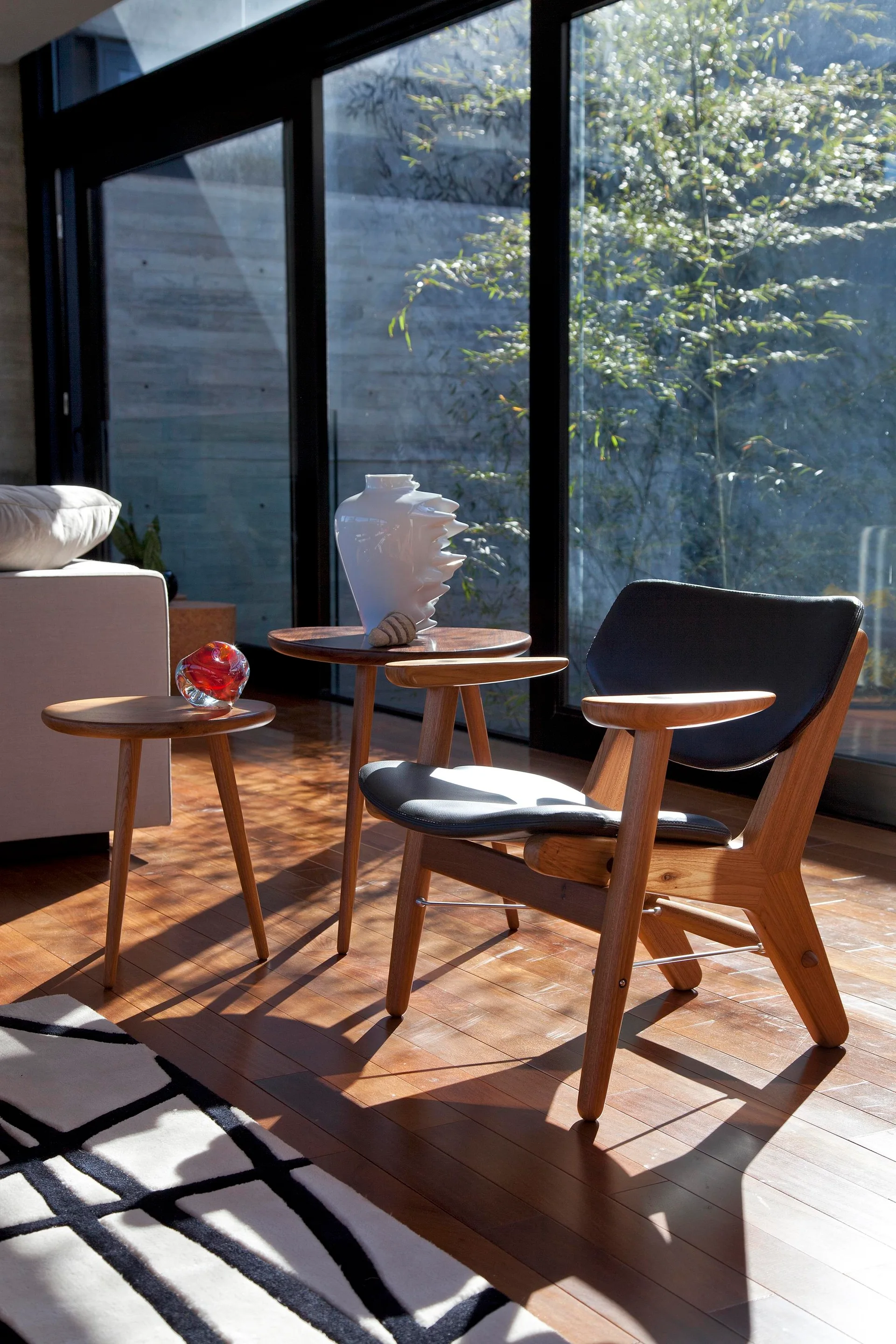 A wooden armchair with black upholstery sits next to two round side tables in a modern sunlit room with large windows.