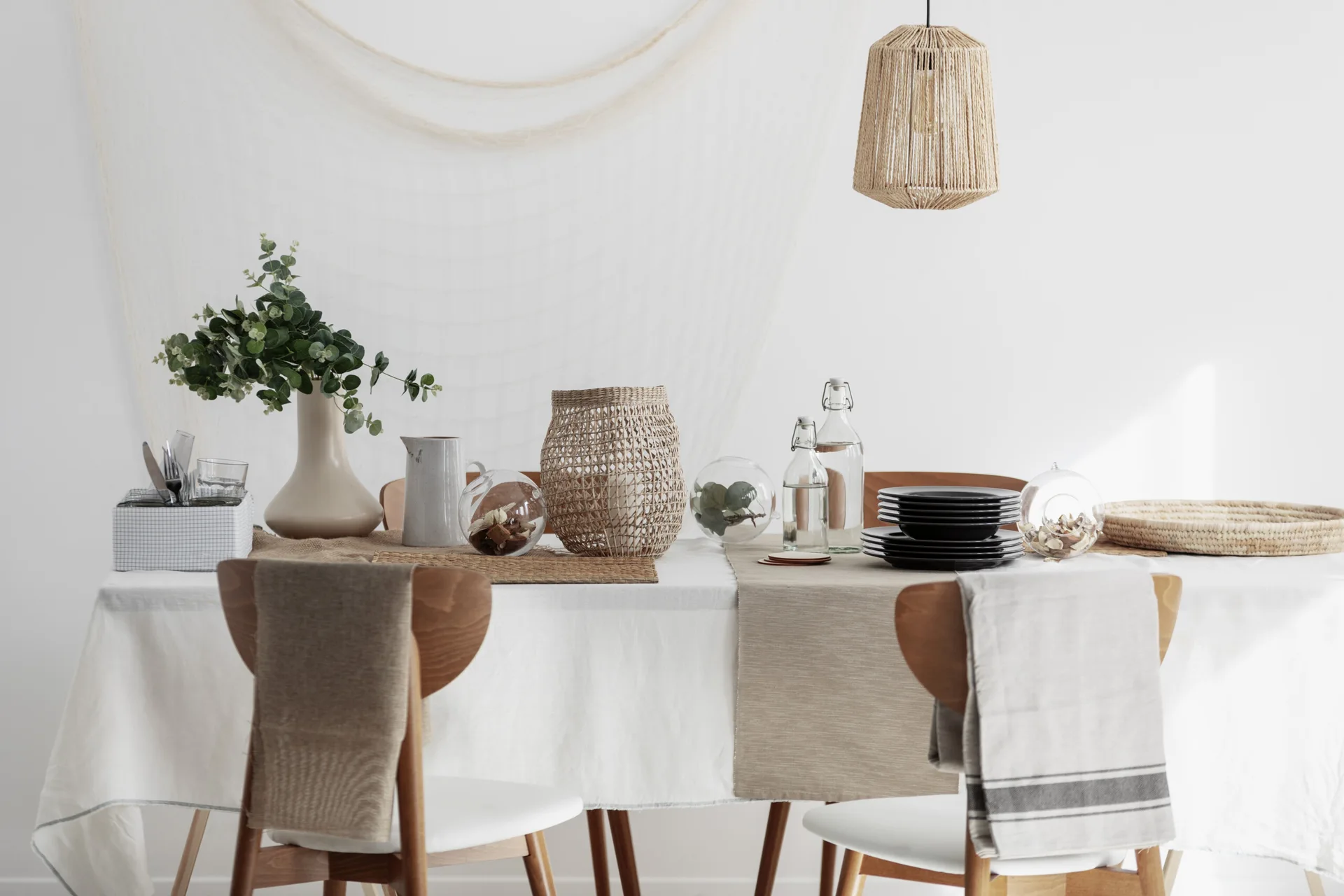 A dining table set with white linens, beige runners, a stack of black plates, eucalyptus in a ceramic vase, and a woven pendant light.