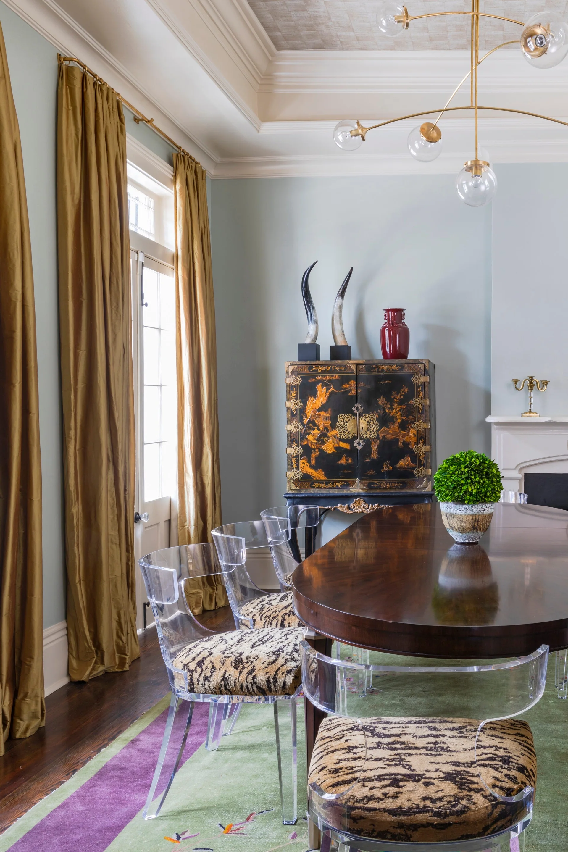 Clear acrylic dining chairs with animal-print cushions surround a dark wood table in a pale blue dining room with gold curtains.