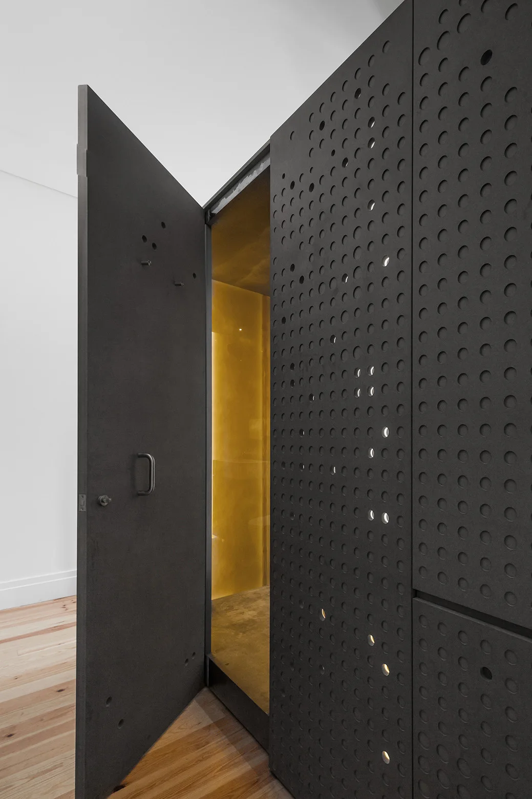 An open matte black door next to a perforated black screen wall reveals a glowing gold metallic interior over light wood flooring.