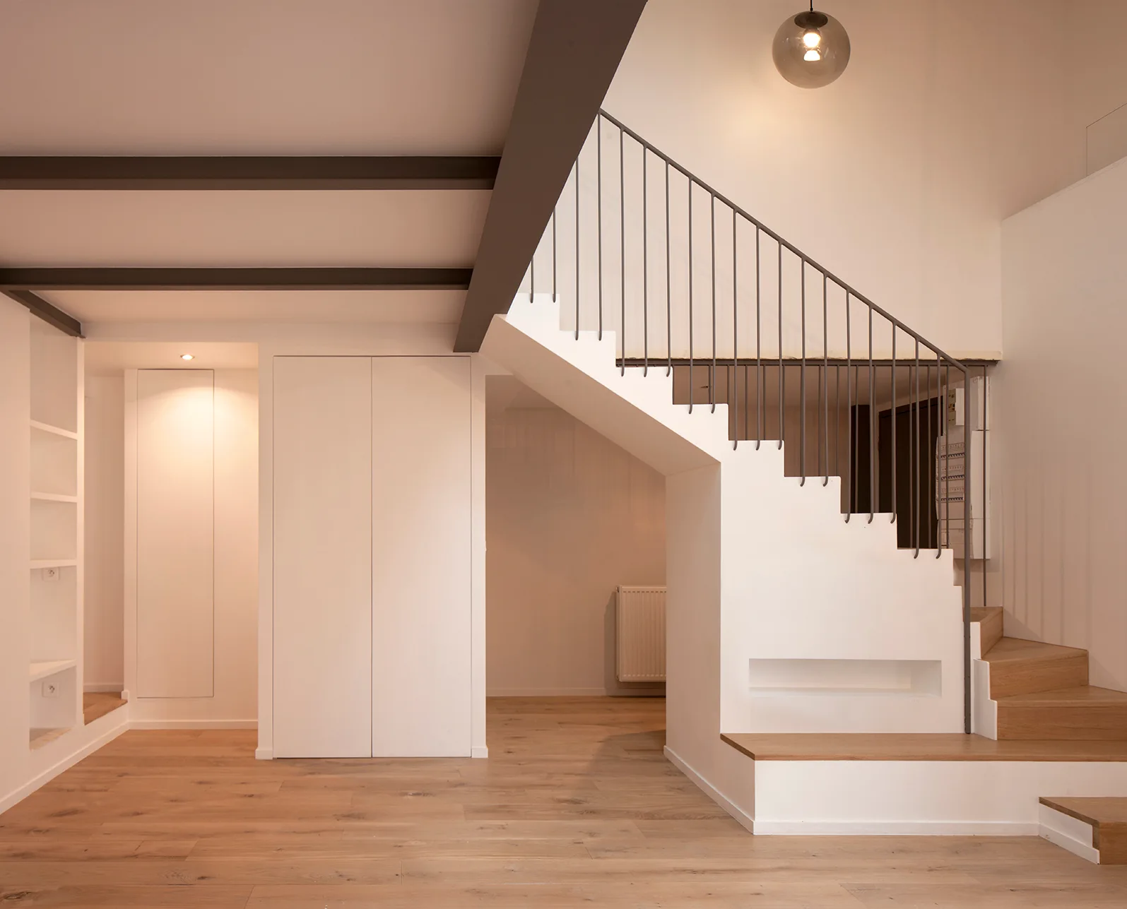 A minimalist entryway featuring a white staircase with light wood treads, thin dark metal railings, and integrated white storage cabinets.