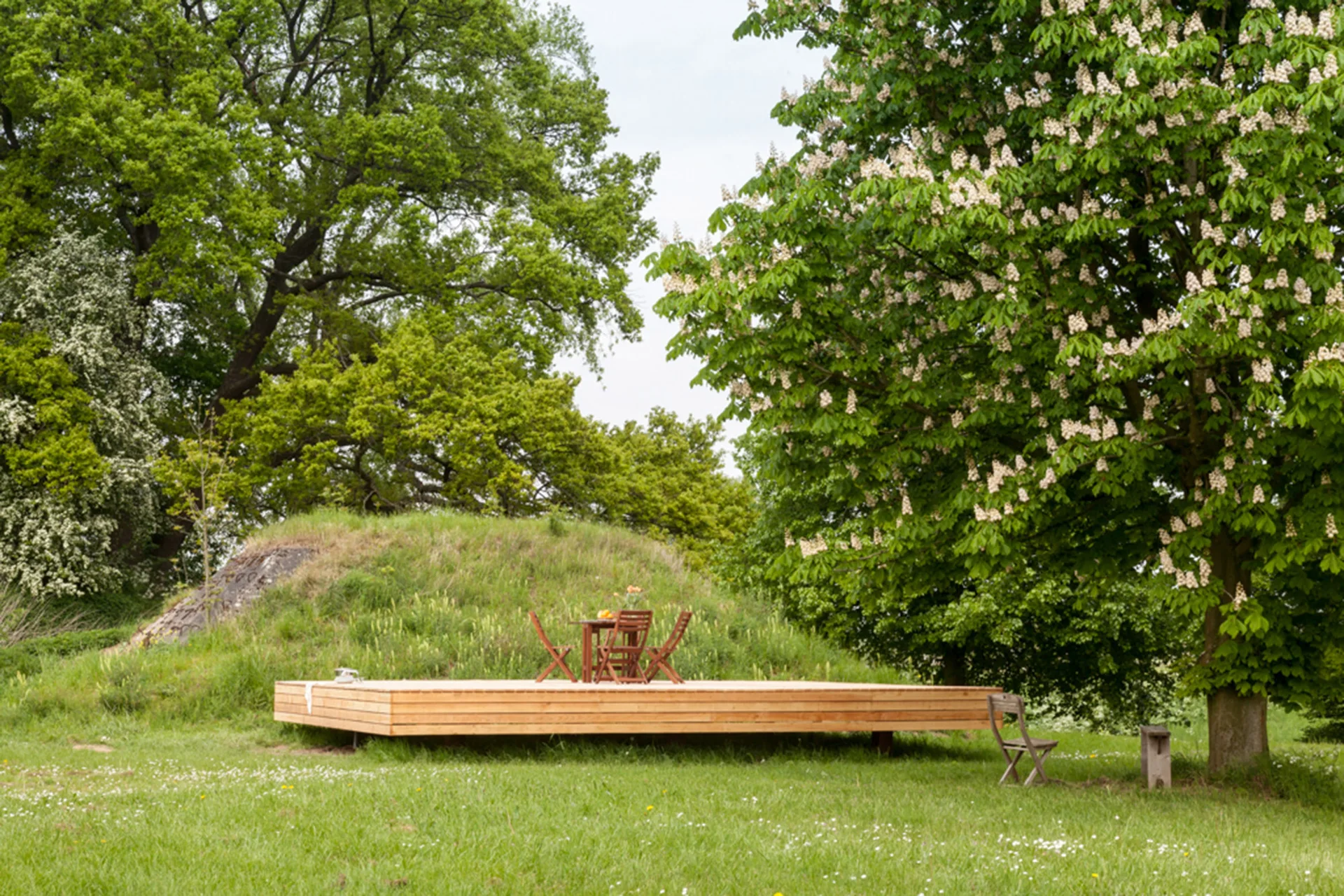 A raised wooden platform deck sits in a grassy meadow with a small wooden dining table and chairs near a large green tree.