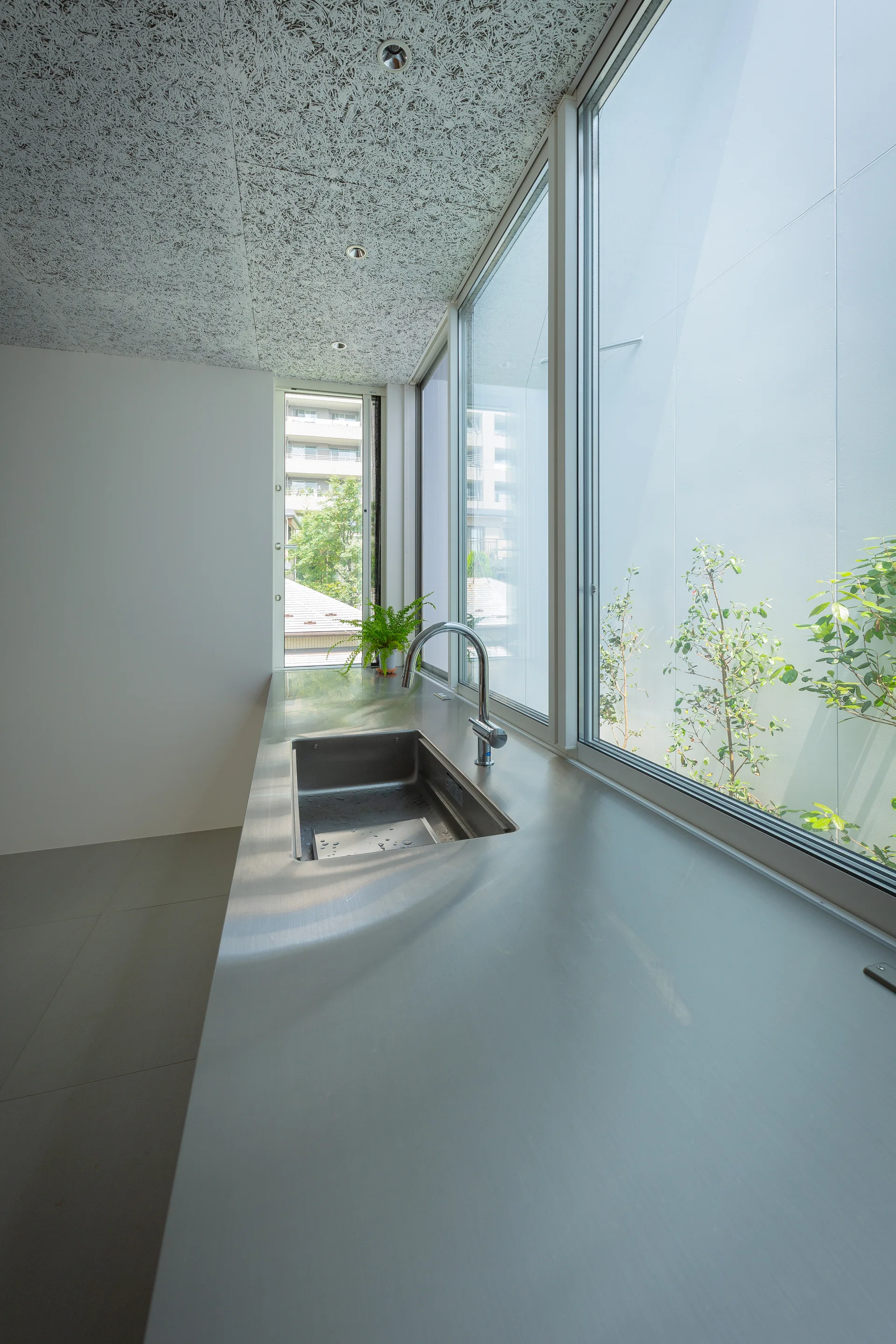 A minimalist kitchen features a long stainless steel countertop with an integrated sink next to a wall of large windows.