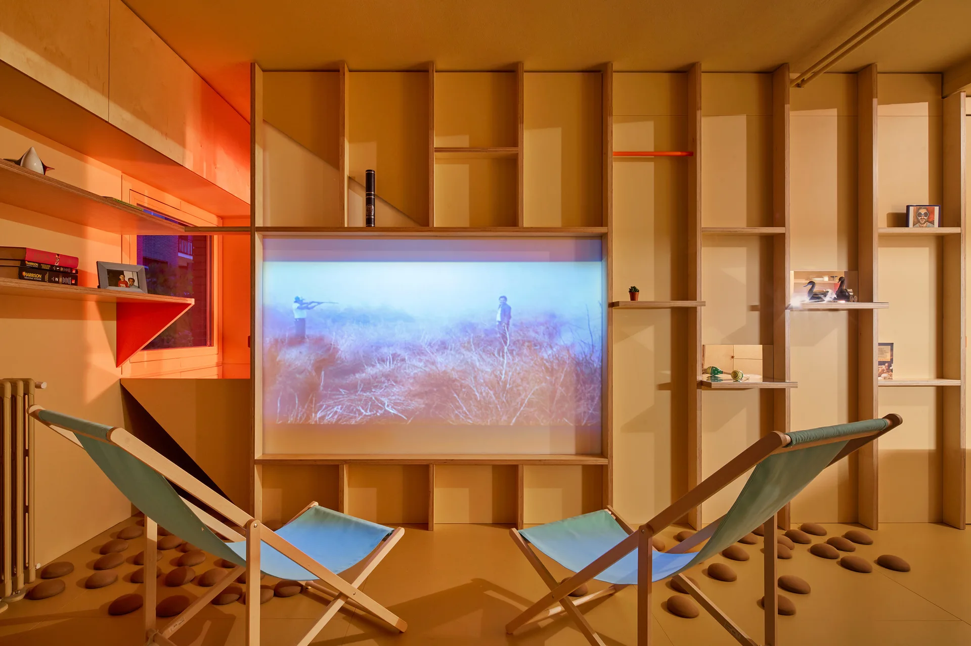 Two blue sling lounge chairs face a projection screen integrated into a custom warm wood shelving unit on an ochre floor.