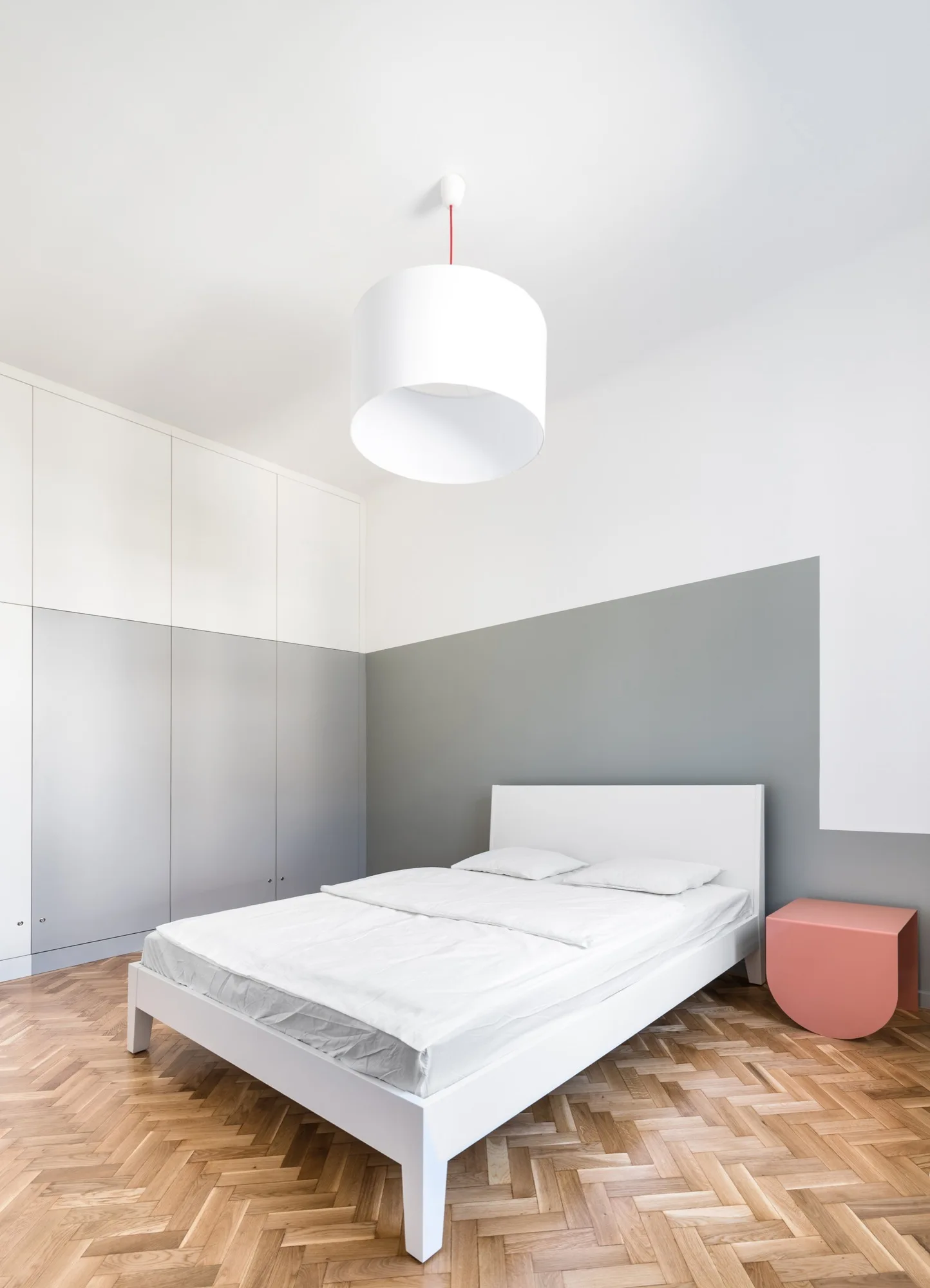 A minimalist bedroom features a white platform bed, a diagonal grey and white wall, built-in wardrobes, and herringbone wood floors.