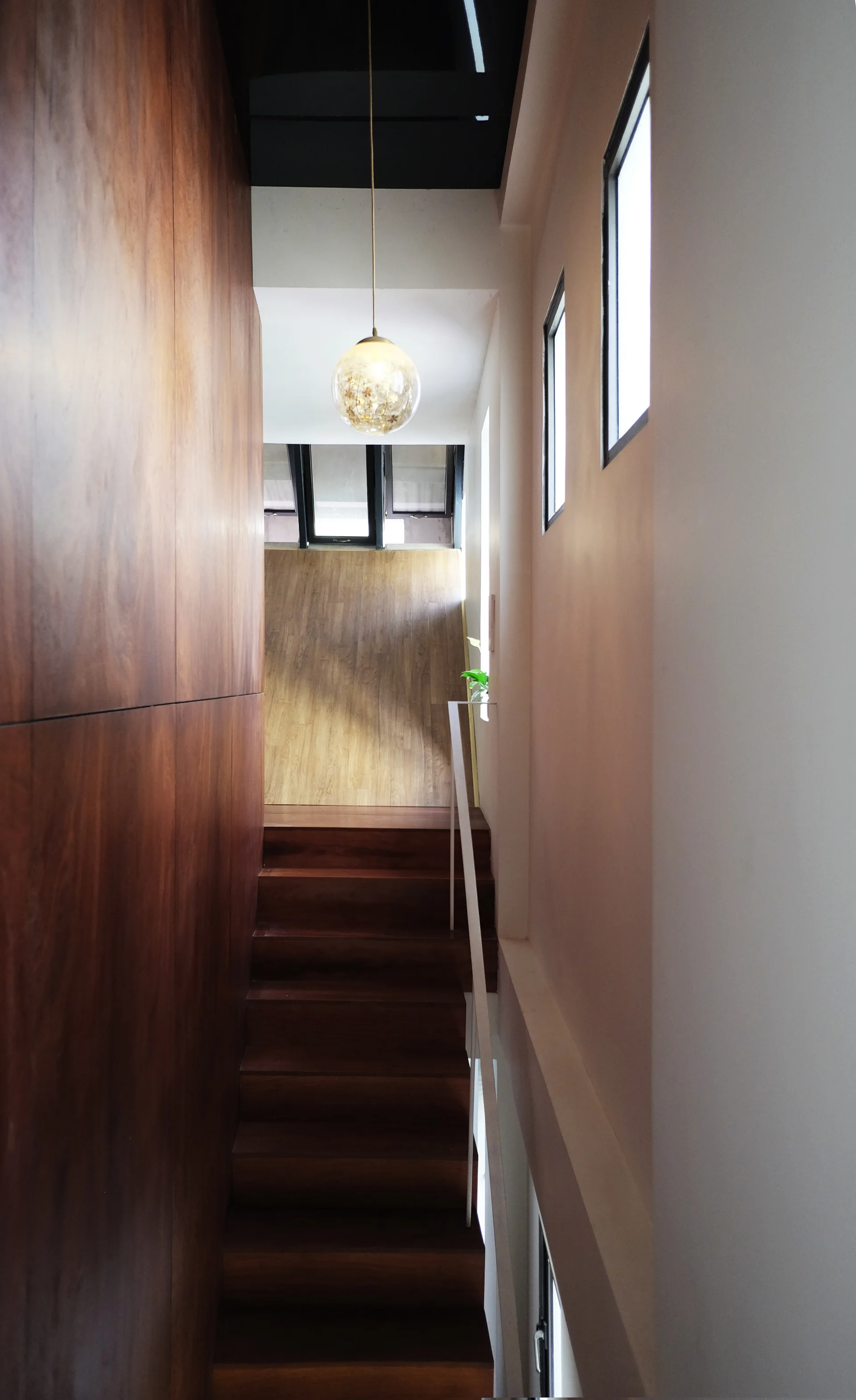 A narrow wooden staircase with dark wood steps and matching wall paneling, featuring a slim white handrail and a glowing globe pendant light.
