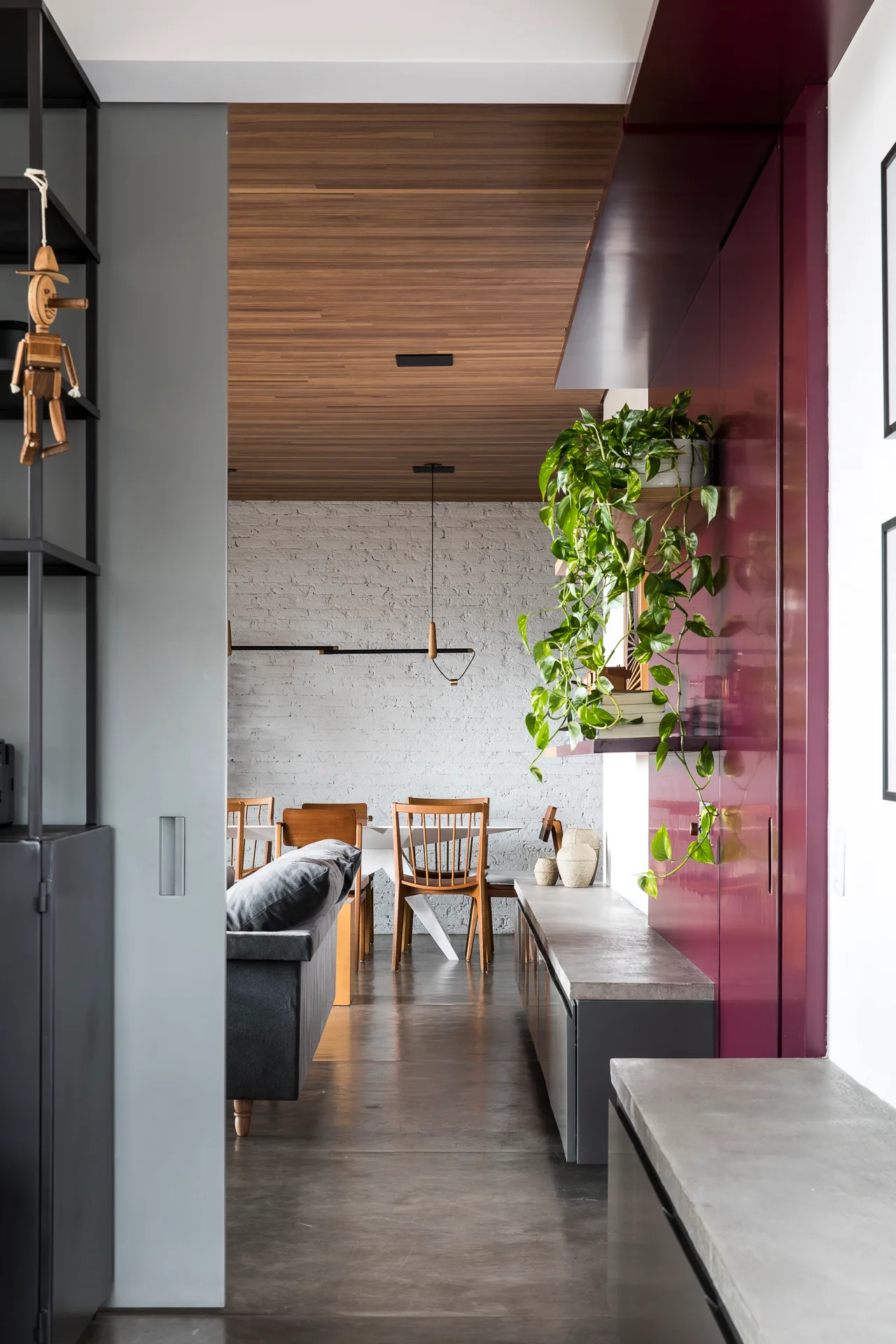 An open dining space features a wood slat ceiling, a painted white brick wall, wooden dining chairs, and a glossy magenta cabinet panel.