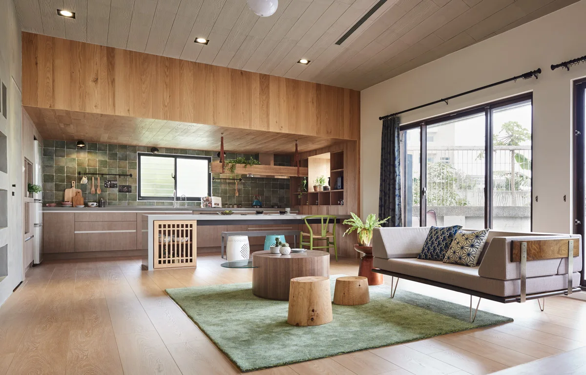 An open-plan kitchen and living room featuring natural wood cabinets, a green tiled backsplash, a light wood coffee table, and a grey sofa on a green rug.