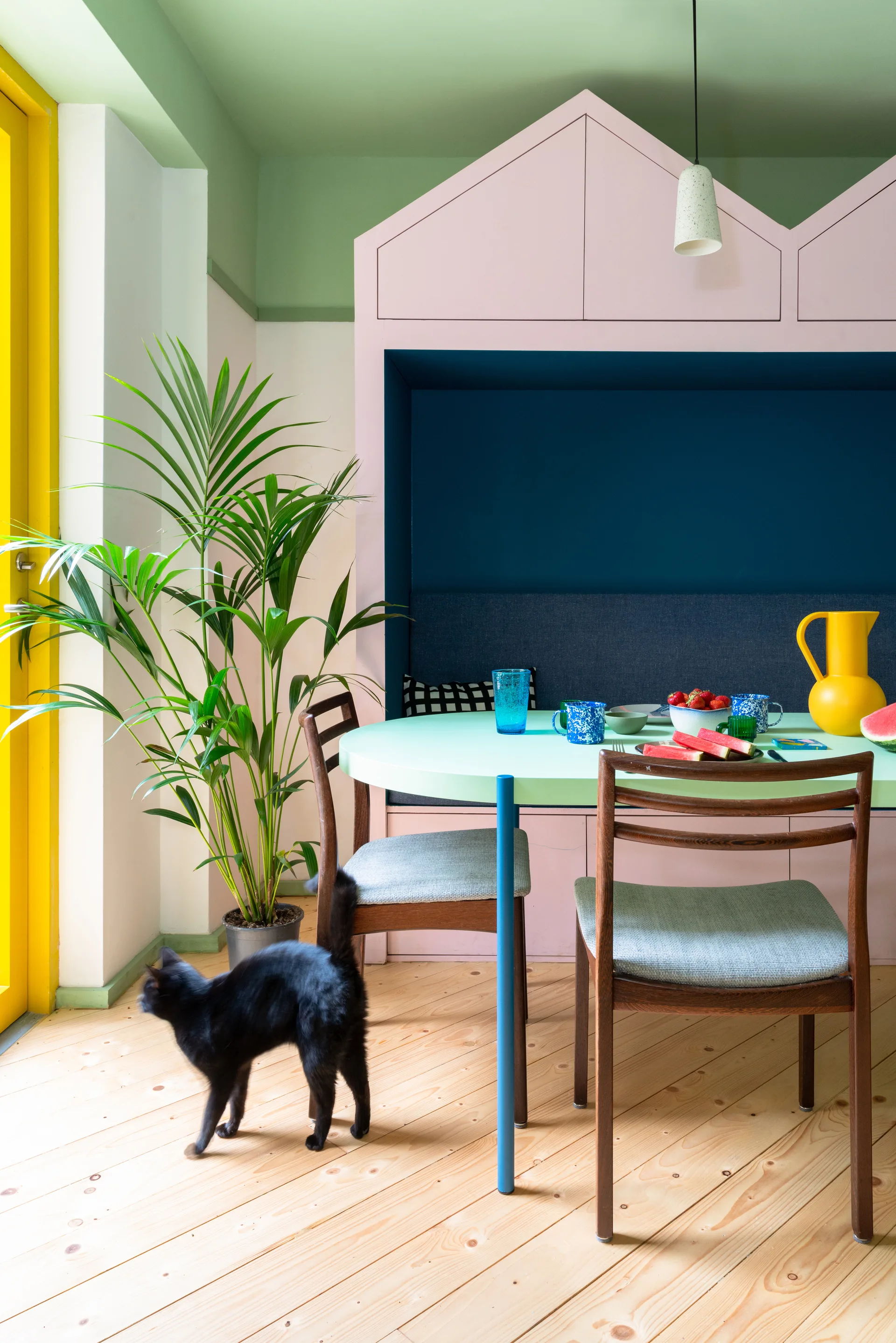A colorful dining area featuring a house-shaped pink built-in bench with a navy cushion, a round mint green table, and wooden chairs.