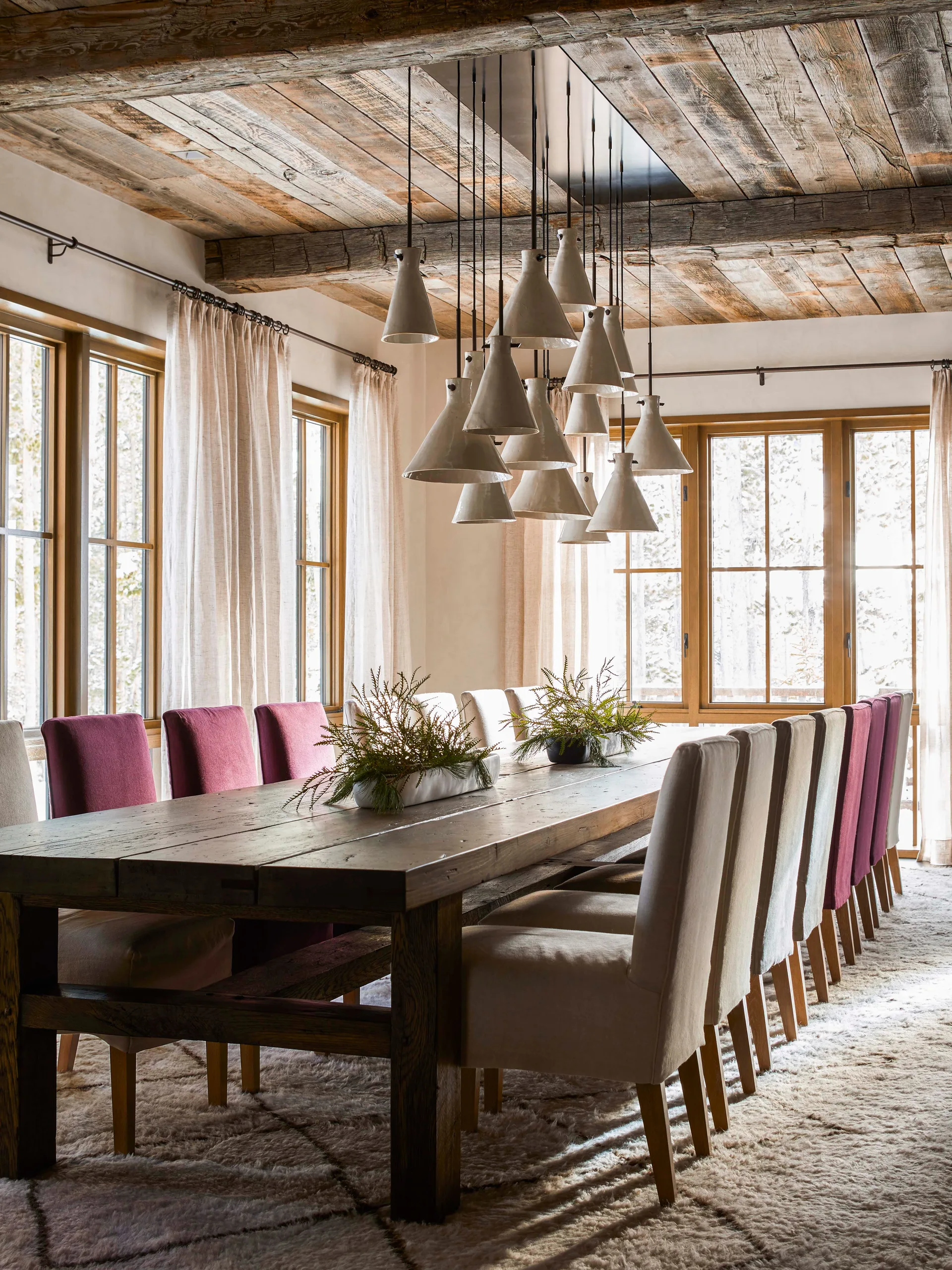 A long wooden dining table surrounded by cream and berry-colored chairs under a large cluster of cone pendant lights and a reclaimed wood ceiling.