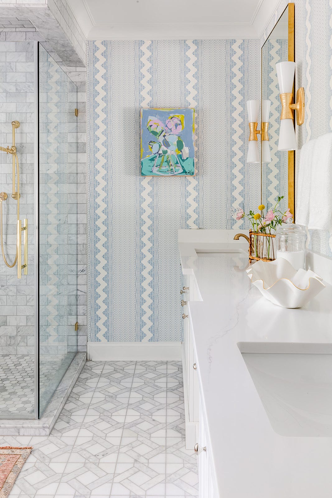 A bathroom vanity features blue striped wallpaper, a white countertop, gold sconces, a gold-framed mirror, and a marble-tiled glass shower.