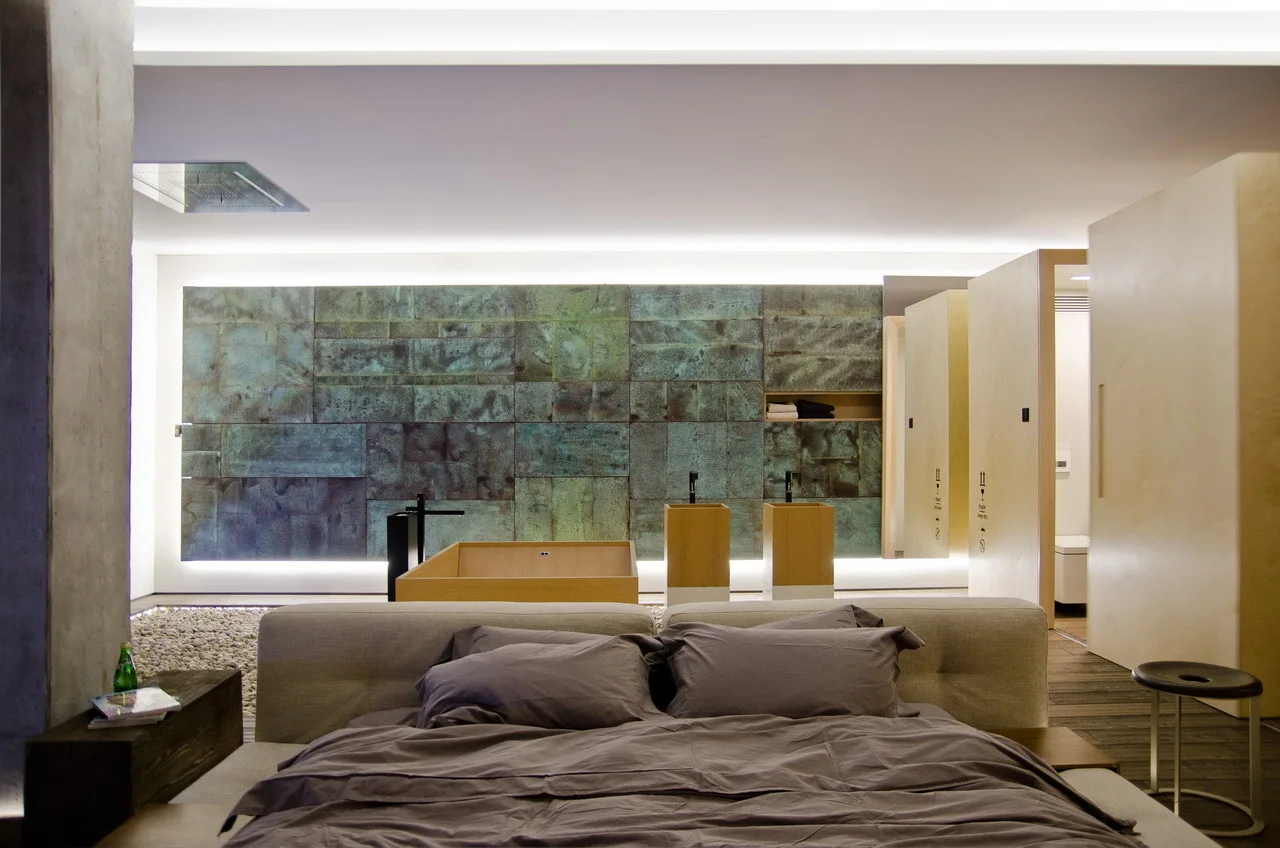 An open-concept bedroom features a low grey bed facing an ensuite bathroom with an oxidized green metal wall, wooden sinks, and a pebble floor boundary.